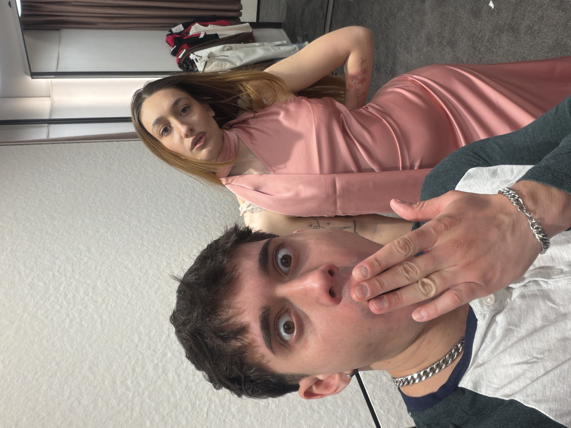 A photograph shows a man and a woman in what appears to be an interior space. The woman is in a light pink satin dress. The man is wearing a white shirt with a dark collar and silver chain. His hand is up near his mouth, with a surprised expression. The location is unknown, and there is no indication this is in Sofia, Bulgaria.
