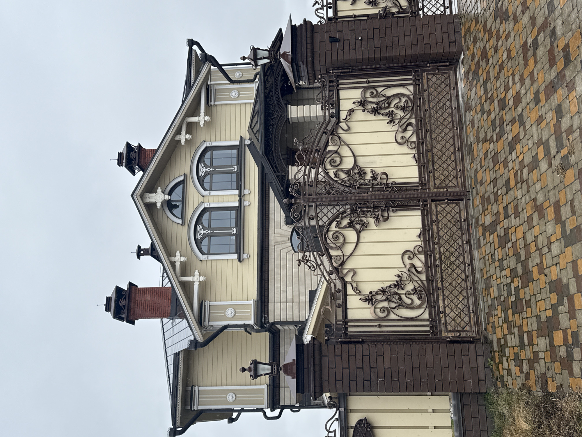 The image captures a house and its elaborate gate, likely located in Sviyazhsk, Russia, based on architectural styles common to the region. The house features a cream-colored facade with intricate woodwork, including ornate window frames and decorative elements along the roofline. It has a steeply pitched roof with dormer windows and a brick chimney. The gate is made of dark metal with a decorative wrought-iron design and brick pillars. The pavement beside the gate is comprised of multicolored bricks. The sky is overcast. No people are visible in the frame.