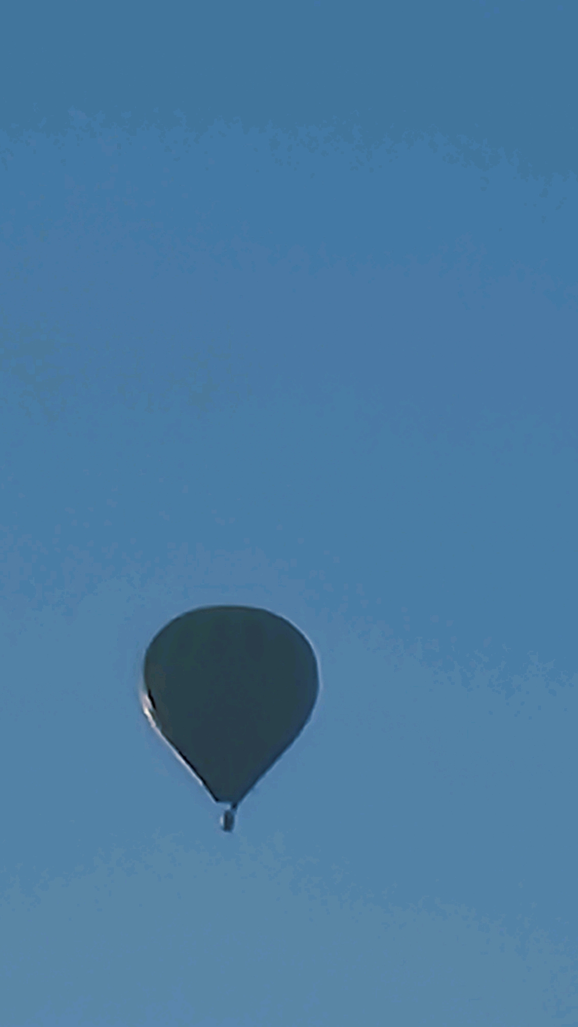 A dark, heart-shaped object, possibly a hot air balloon or drone, is suspended in a clear blue sky. The object is located in the sky above Sant Joan Les Fonts, Spain.