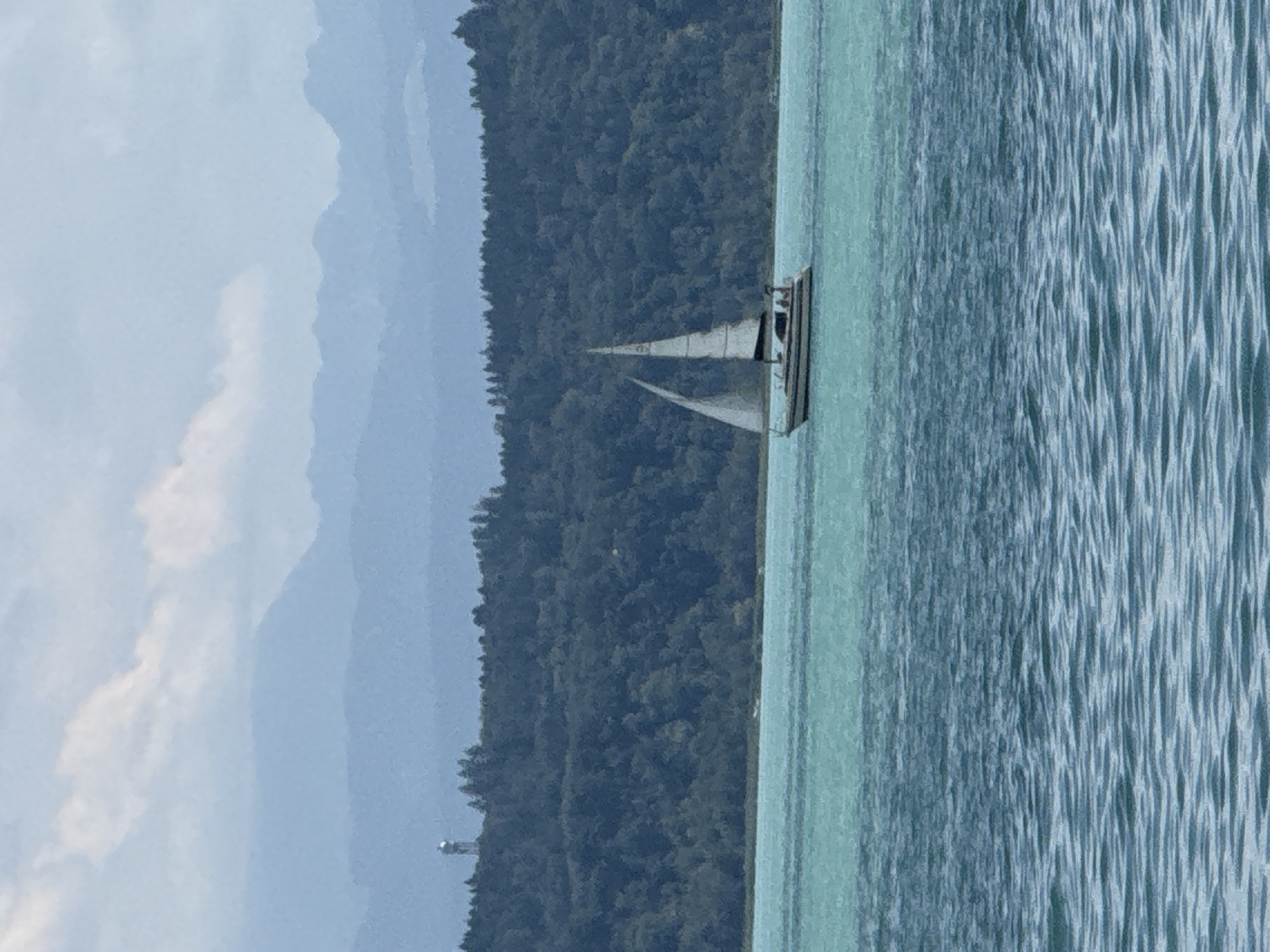 A sailboat navigates on a body of water, with a tree-covered hillside in the background. The sky is overcast with light clouds, and distant hazy mountains are visible. A small tower-like structure can be seen on the hilltop. The water is a bright turquoise color, with ripples on the surface. The sailboat has two masts with sails and appears to have at least two people on board. The scene is likely a lake or a large bay. This image is a photograph taken in the city of Neuchâtel, Switzerland, possibly overlooking Lake Neuchâtel.
