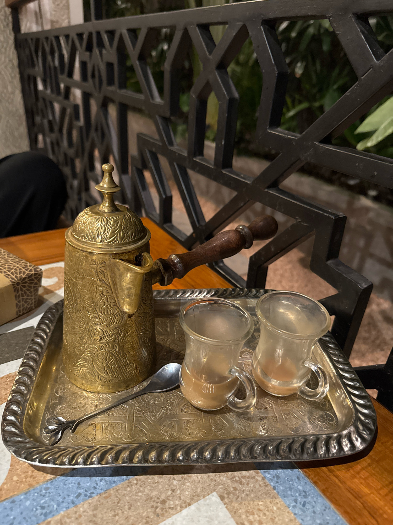 A tarnished, ornate brass coffee pot with a wooden handle and a decorative lid sits on a decorative silver tray. The tray also holds two small, clear glass cups filled with a light brown liquid, and a single silver spoon with a decorative handle. The tray rests on a wooden table, with a patterned box partially visible to the left. In the background, a black metal decorative fence with geometric patterns separates the foreground from lush green foliage. The scene appears to be set outdoors on a patio or balcony, possibly at a cafe or restaurant in Denpasar, Indonesia. A person's dark clothing is visible to the left of the coffee pot.