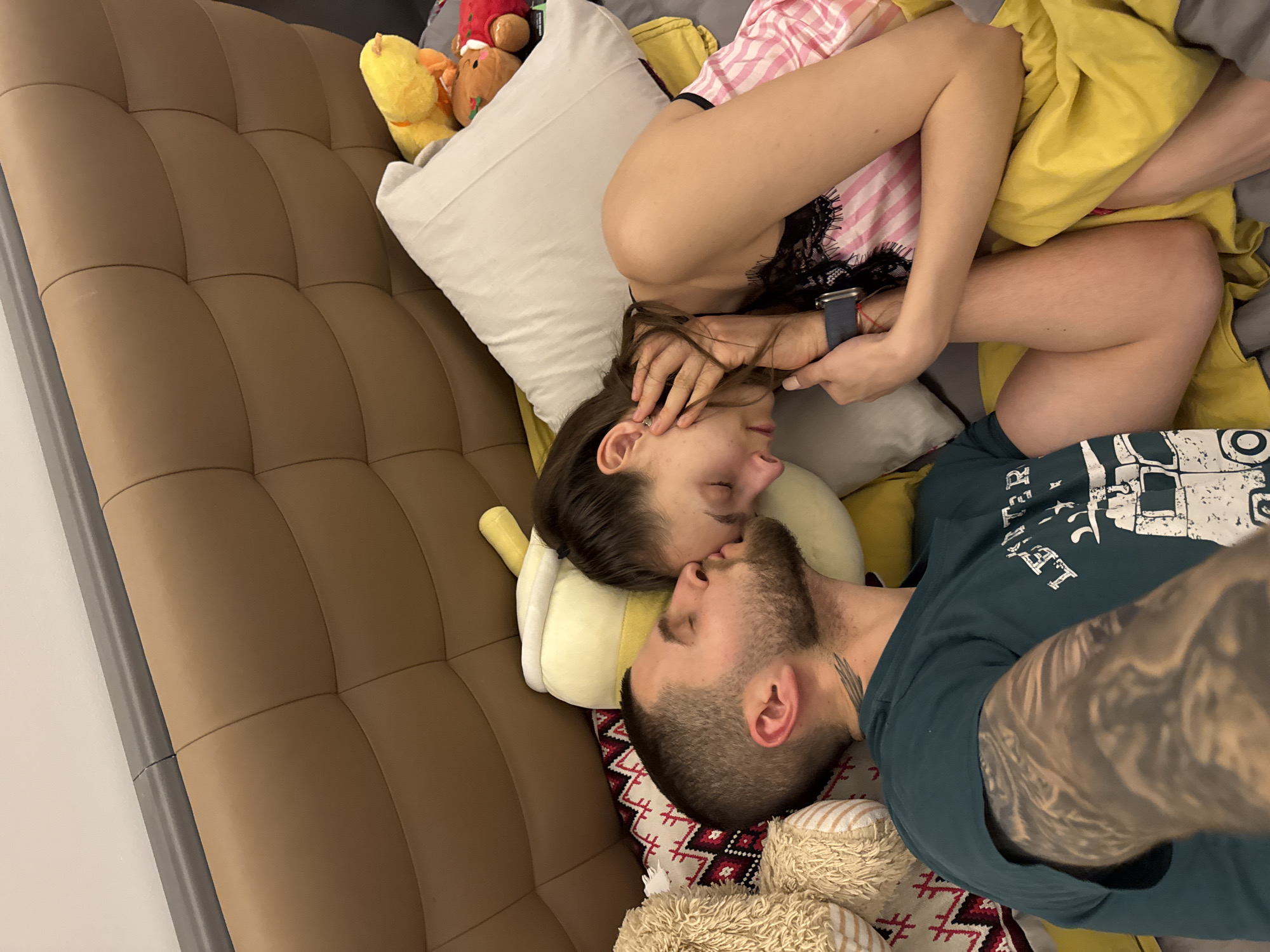 A man and a woman are lying on a brown tufted sofa, possibly in Sofia, Bulgaria, based on visual cues and the lack of contradictory information. The woman, wearing a pink striped top with black lace trim, is lying on her side with her head resting on a white pillow. Her left hand is near her face, and she is wearing a smartwatch on her left wrist. Her legs are bent and covered by a yellow blanket. The man is lying next to her, also on his side, with his face close to hers. He has short hair and a beard, and he is wearing a dark green t-shirt. A tattoo is visible on his left arm and on his neck. Several plush toys, including a yellow duck and a brown bear with Christmas decorations, are arranged around them on the sofa. A red and white patterned pillow is visible beneath them. The sofa is placed against a light-colored wall.