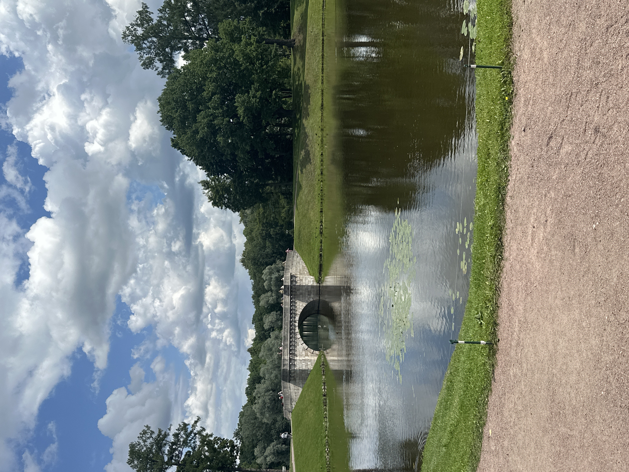 The image captures a view of the park in Gatchina, Russia, featuring an arched stone bridge spanning across a calm body of water. The bridge, constructed from light-colored stone, has decorative elements along its top and sides, and several people are visible walking on its surface. The water reflects the bridge and the surrounding lush greenery, with lily pads dotting its surface. The park's landscape includes well-maintained grassy slopes leading down to the water's edge, with a gravel path in the foreground. A line of small white and green posts delineates the edge of the grassy area. In the background, mature trees form a dense forest line, and the sky is filled with a mix of blue patches and white, puffy clouds. The overall scene depicts a serene park setting on a bright, cloudy day.