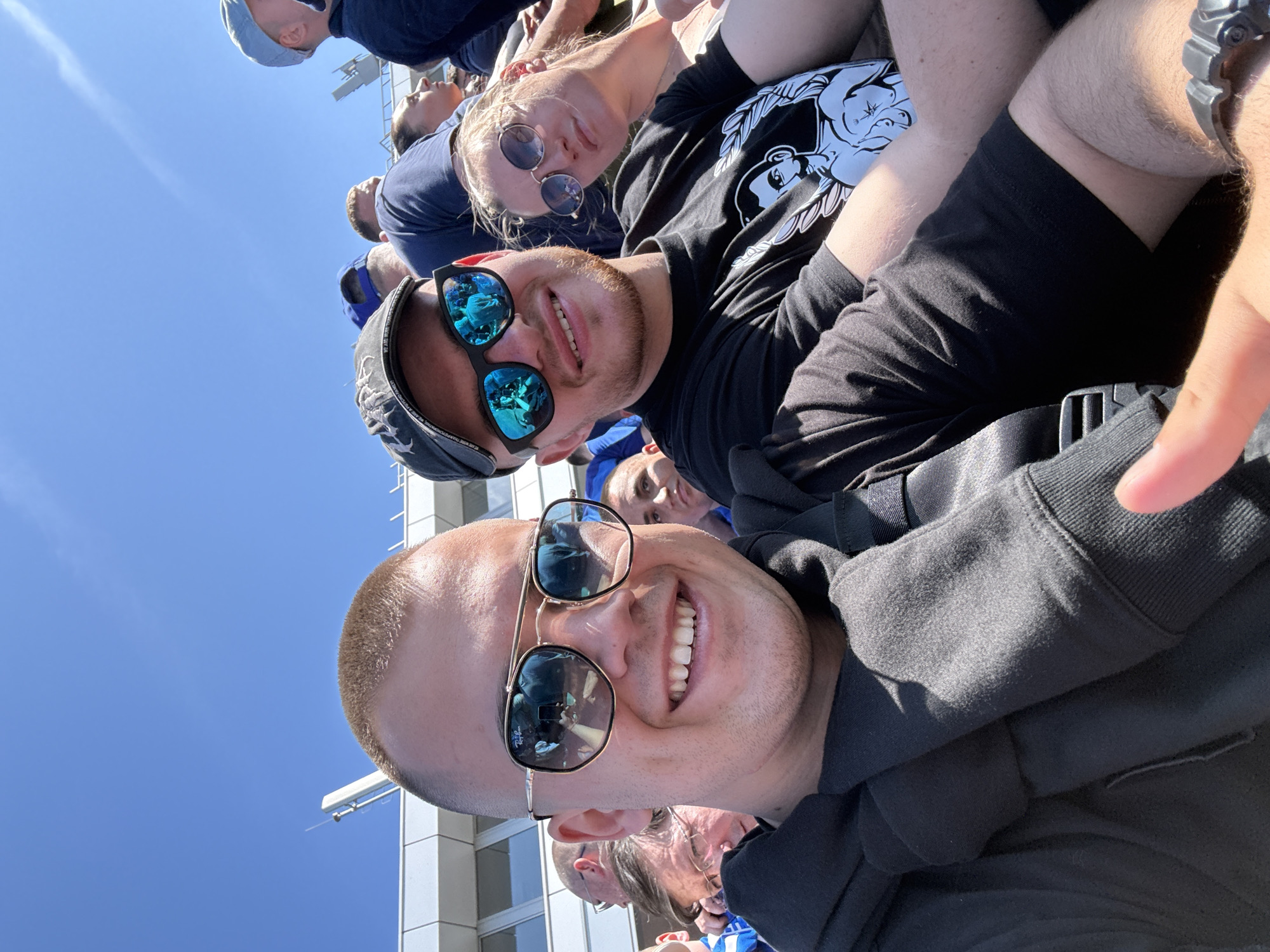 The image shows a selfie taken by a person wearing gold-rimmed sunglasses and a black hooded sweatshirt. The reflection in the sunglasses shows a person holding a phone, presumably taking the picture. Behind the first person is another person wearing sunglasses and a baseball cap, smiling. A third person with round sunglasses is visible behind them. All individuals are likely attending an event, as other people are visible in the background. The sky is blue, and a building is partly visible, suggesting an outdoor location in a city. The specific city is not identifiable.