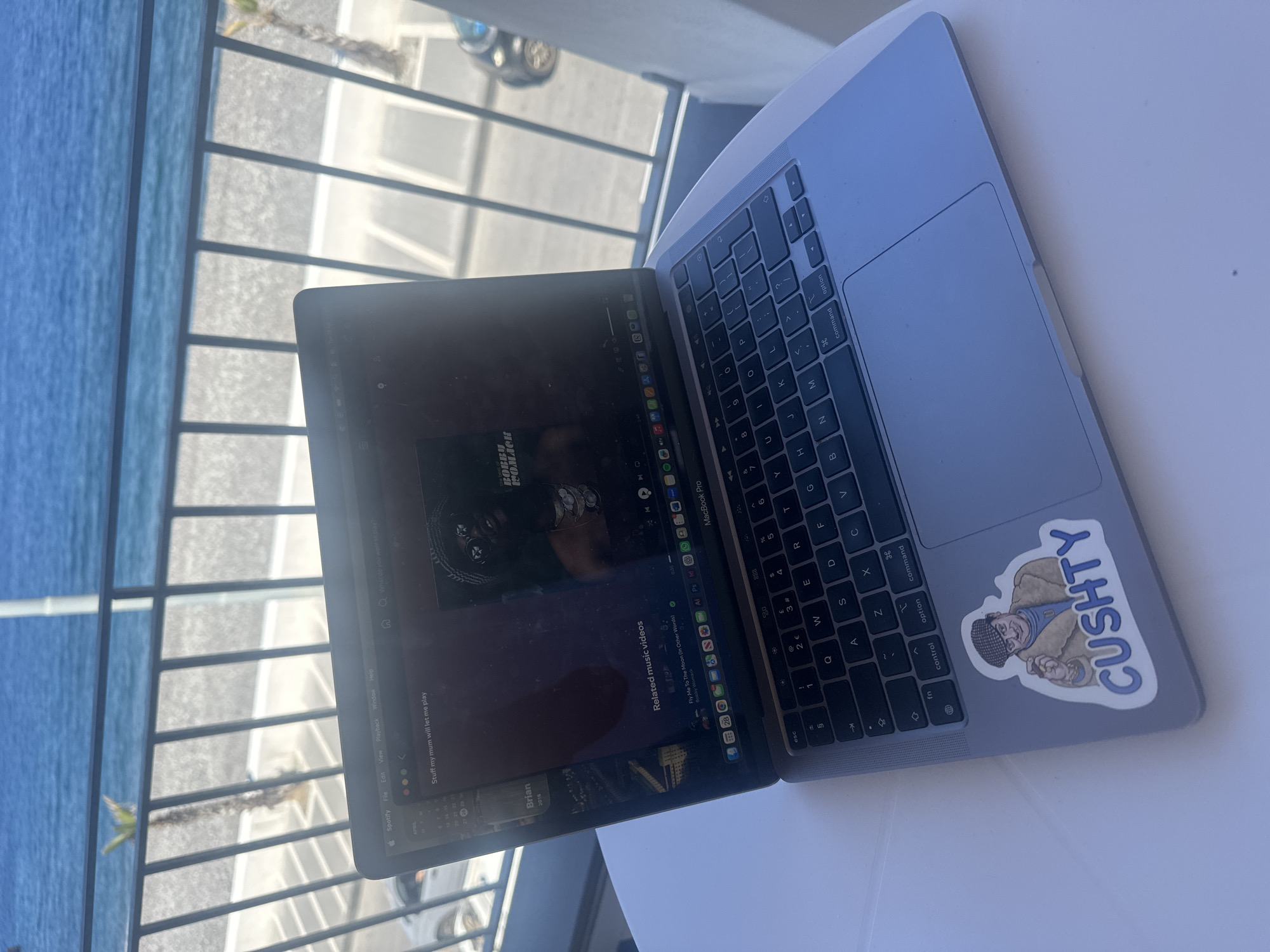 The image displays a silver Apple MacBook Pro laptop placed on a white table, open and positioned beside a window or balcony with a view of the sea. Bright sunlight illuminates the scene, reflecting off the blue water to the left. Beyond a balcony railing, a blurred background reveals pavement, what appears to be a palm tree, and a partial view of a car, indicative of a coastal location like Lipari, Italy.
The laptop screen shows a dark interface, resembling a music or video streaming application. The prominent display features an image of a dark-skinned man with braids or dreadlocks and sunglasses, labeled "BOBBY WOMACK." Below this, a section for "Related music videos" highlights "Fly Me To The Moon (In Other Words)" by Bobby Womack. Visible on-screen text includes "Spotify," standard application menus like "File," "Edit," "View," "Playback," "Window," and "Help," along with a search bar reading "Q What do you want to say?". A sidebar contains the phrase "Stuff my mum will let me play" and a calendar displaying dates for April. The dock at the bottom of the screen shows various app icons.
A notable detail is a "CUSHTY" sticker on the laptop's right palm rest, depicting a cartoon character wearing a flat cap. The laptop's QWERTY keyboard is visible, with keys including "esc," "command," "option," "control," and "fn." The scene suggests a daytime, leisure setting, possibly a vacation, where the user is engaged with digital entertainment while enjoying an outdoor view.