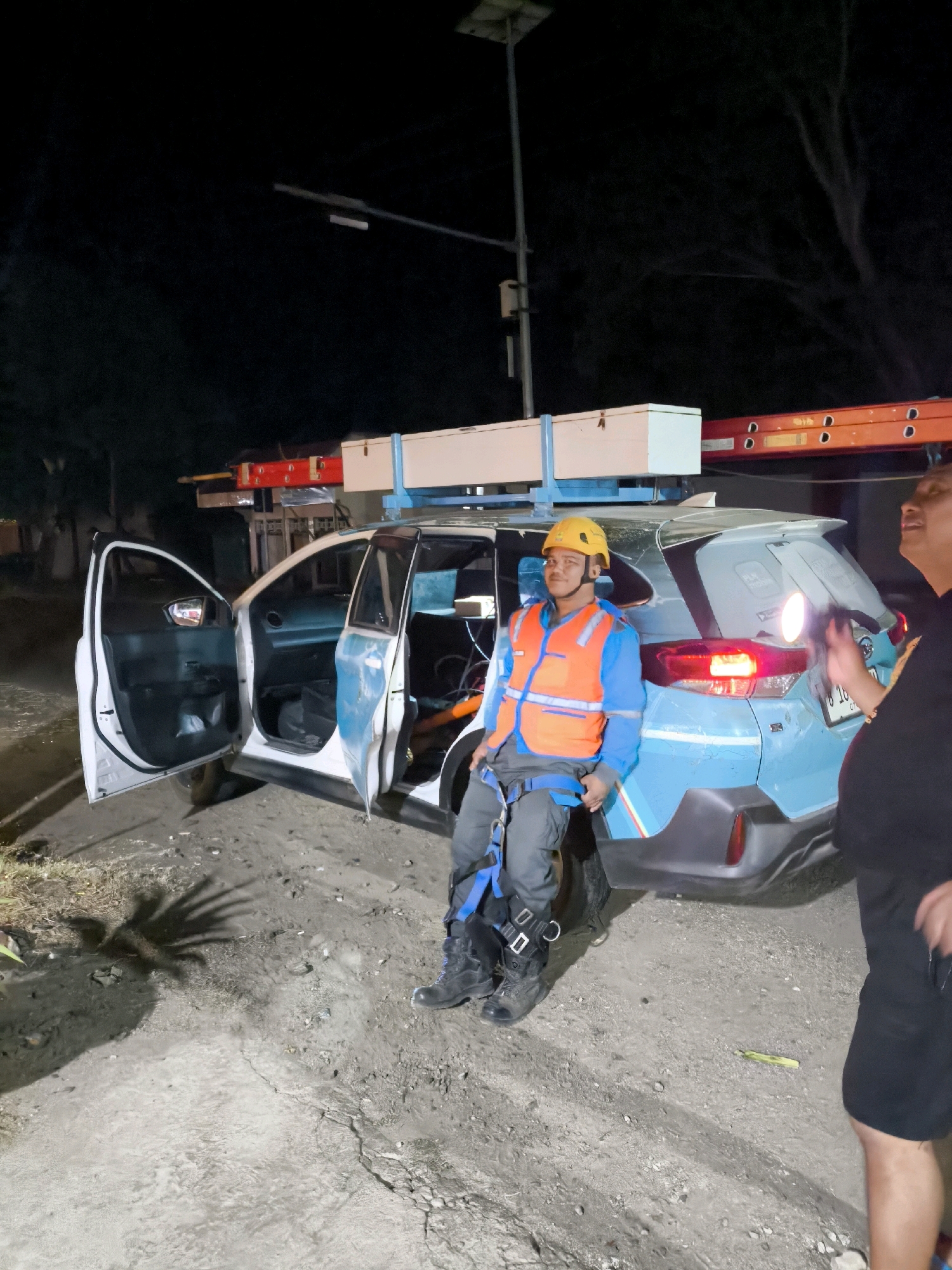 In an outdoor, unpaved area at night, a light blue SUV with its doors open is parked. A man in a yellow hard hat, orange safety vest, and a harness is seated in the driver's seat area. A ladder is secured to the roof rack of the SUV. To the right of the SUV, another person wearing a black shirt and shorts is partially visible, holding a flashlight. The ground is covered in dirt and gravel, with some sparse vegetation and debris. A palm tree shadow is cast on the ground. The scene appears to be in an undeveloped area on the outskirts of a city in Indonesia.