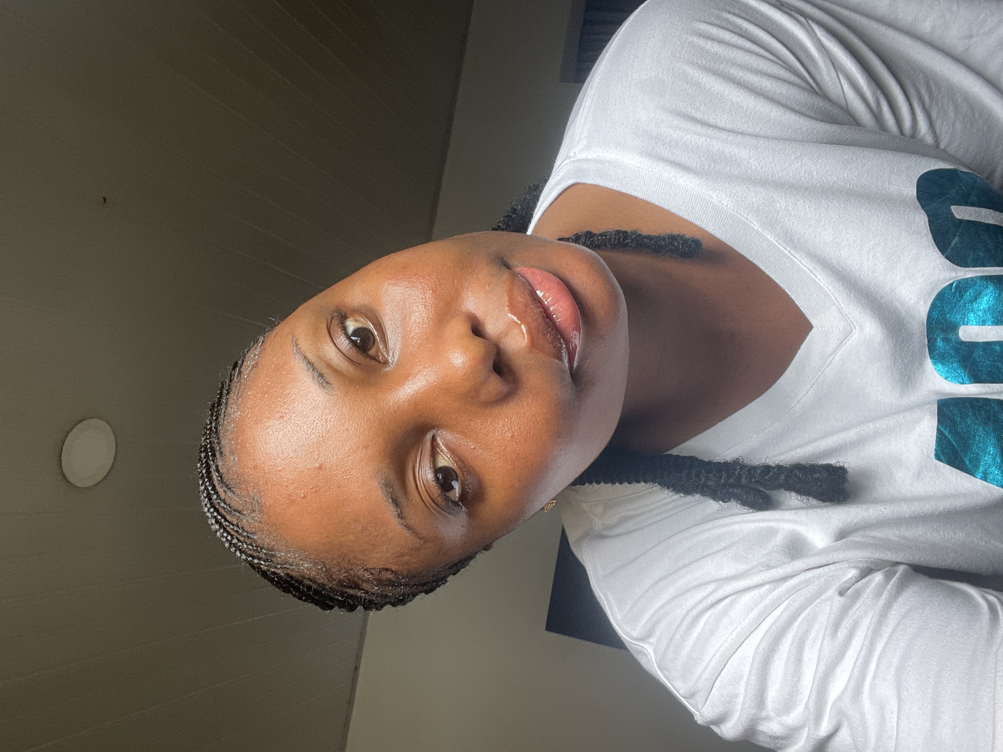 A close-up, slightly tilted portrait features a Black woman with dark skin and cornrow-style braids. The woman is wearing a white V-neck shirt with a teal-colored graphic on the right side of the frame. Her lips are glossy, and her eyes are light brown. The background shows a ceiling with a light fixture, and the top of a dark-colored object is visible near the woman's right shoulder. The location cannot be determined based on the image.