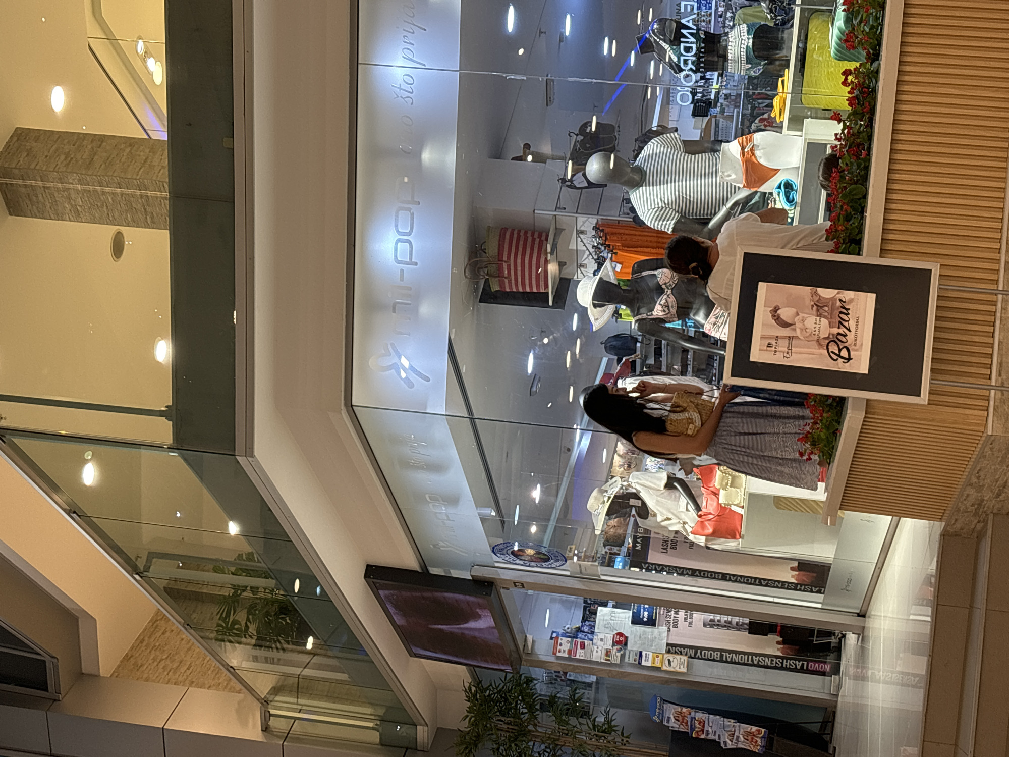 The image shows the exterior of a boutique store in Budva, Montenegro. The store's name, "mi-pop", is visible on the glass facade. Inside, mannequins are displayed wearing swimwear and casual clothing. A woman is standing near the entrance, looking at the display. In the foreground, a framed poster with the word "Bazar" is placed on a wooden counter. The store appears to be located in a shopping mall, with other shops and escalators visible in the background. The lighting suggests it is either evening or the store is dimly lit.