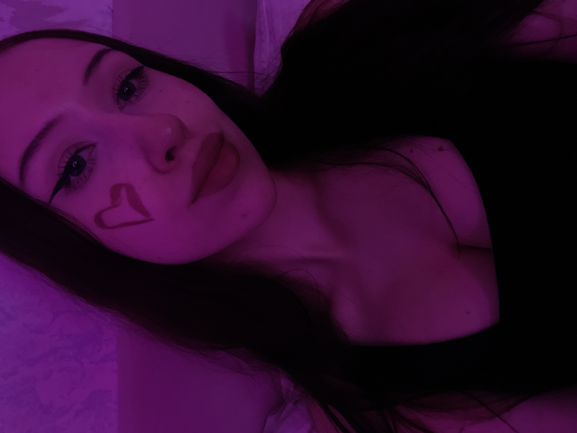 The image shows a close-up, low-angle selfie of a young woman with dark hair, taken indoors. She is wearing a black top and has eyeliner and a heart shape drawn on her cheek. The lighting is predominantly pink, creating a vibrant and atmospheric effect. The background appears to be a textured wall or headboard. There is no information in the image to suggest a location in Votkinsk, Russia.
