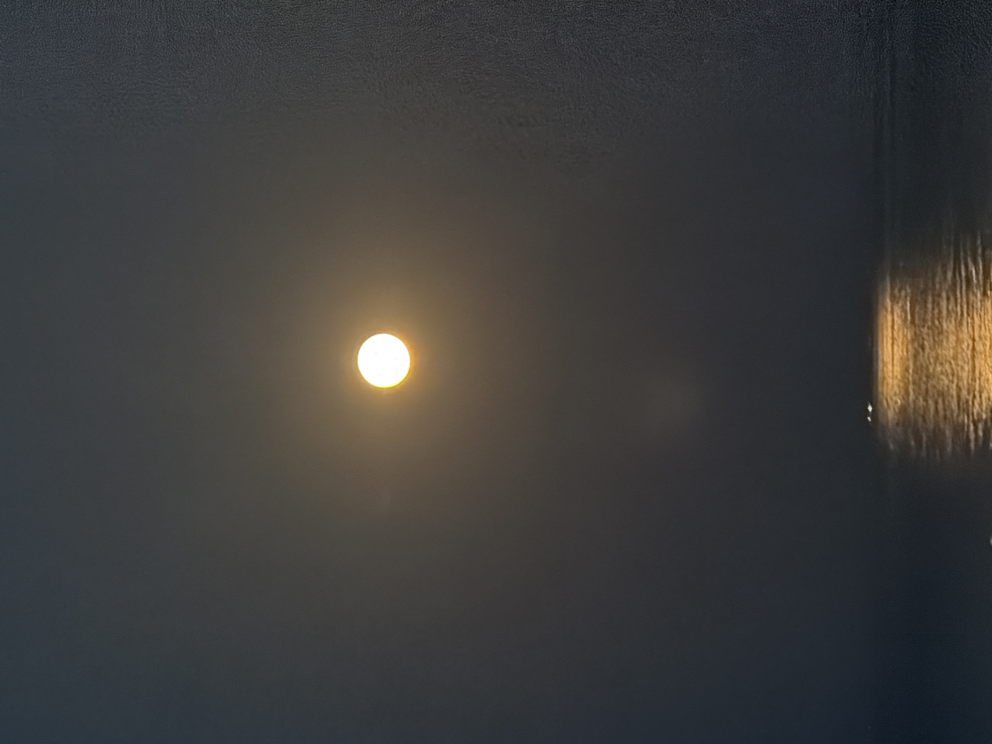 A photograph taken at night in Νέα Καλλικράτεια, Greece, depicts a moonlit scene over a body of water. The moon, a bright, luminous orb, is positioned centrally in the upper portion of the frame, casting a yellowish glow. Its light reflects onto the water below, creating a shimmering, golden path. In the distance, two small, bright lights are visible on the water, likely from a boat or buoy. The overall atmosphere is one of serene darkness, with the moon and its reflection being the primary sources of illumination. The water appears calm, with gentle ripples visible in the reflected moonlight. The sky is dark and hazy, with a subtle texture.