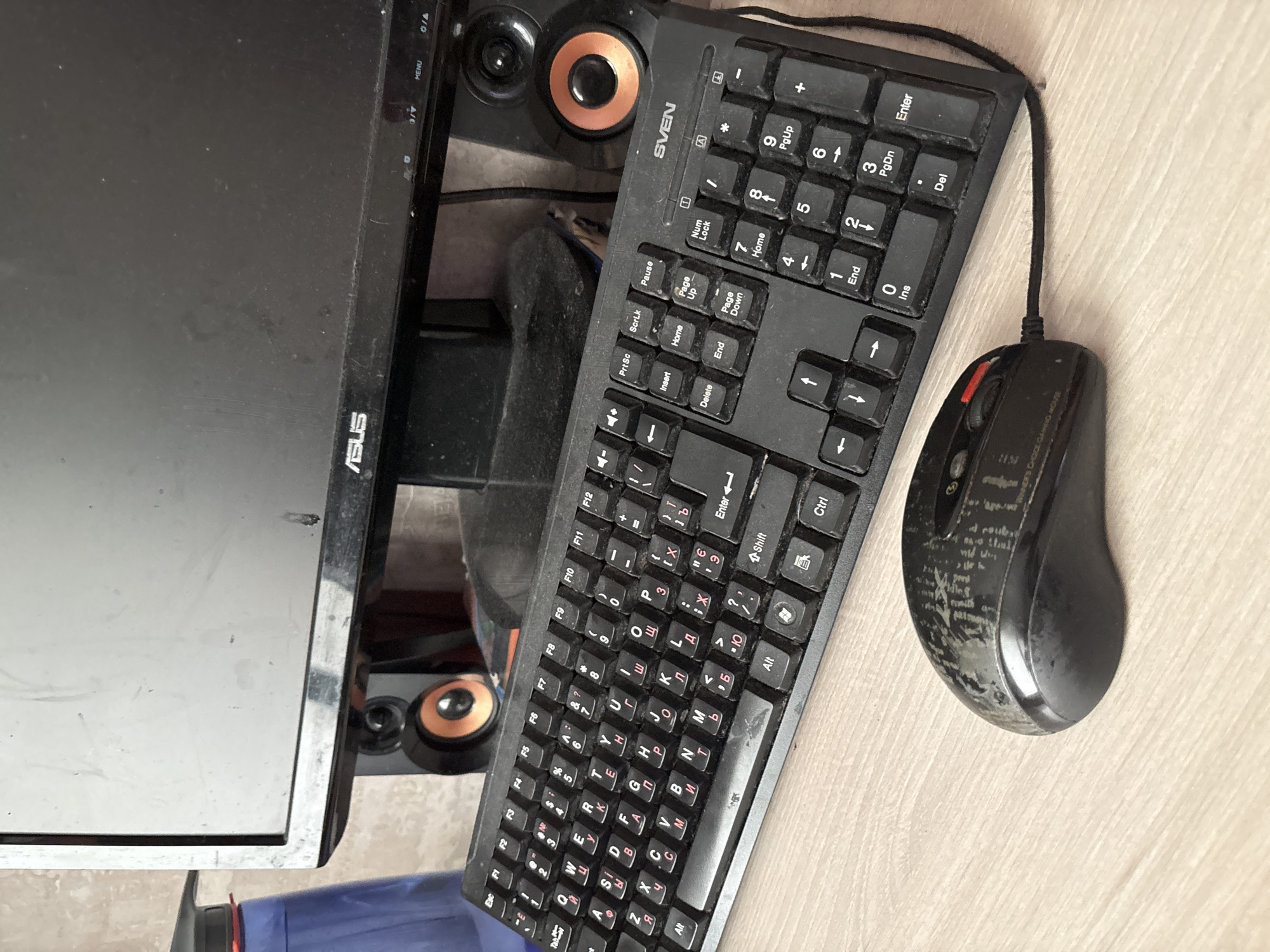 The image shows a close-up of a dusty computer setup on a wooden desk. The setup includes a black ASUS monitor, two small black speakers with orange accents, a black keyboard with both Latin and Cyrillic characters, and a black wired mouse with a red scroll wheel. The keyboard shows signs of heavy use and dust accumulation. The mouse has a worn, patterned surface. In the background, part of a blue bottle with a graduation cap is visible. The scene is indoors, likely in a domestic or office setting. There is no indication of the city of Sochi, Russia, or any people present in the image.