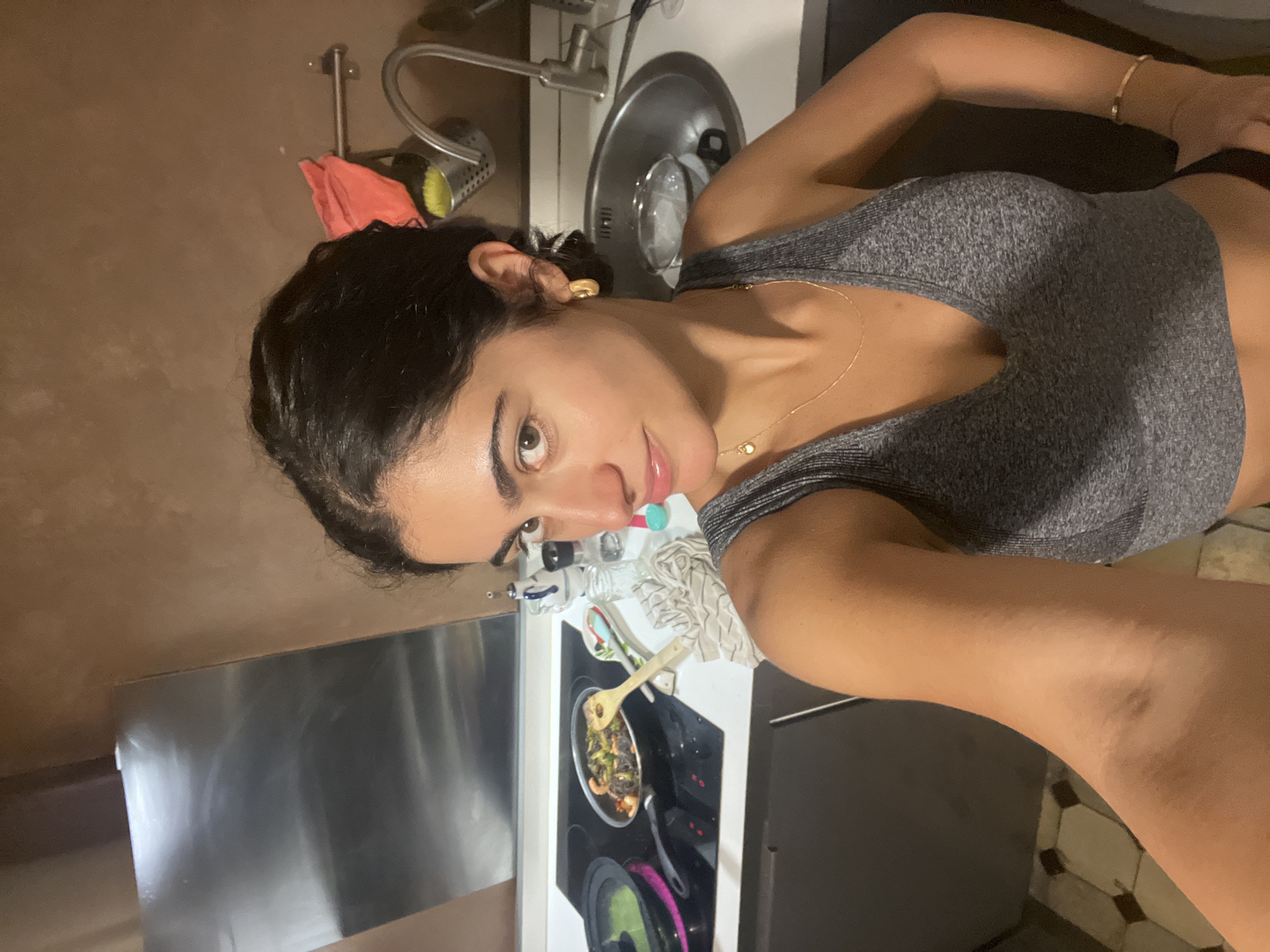The image displays a woman taking a selfie in a home kitchen environment. She has dark hair, dark eyes, and a medium skin tone, looking directly at the camera with a neutral to slightly smiling expression. She is wearing a grey ribbed sports bra or crop top. Accessories include a thin gold chain necklace with a small pendant, a gold hoop earring on her visible left ear, and a thin gold-colored bracelet on her left wrist.
The background details a functional kitchen. To the woman's left, a stainless steel appliance, possibly a refrigerator or dishwasher, is visible. Behind her, a kitchen sink with a faucet is present, alongside cleaning supplies such as yellow and reddish-orange sponges. To her right, on a white countertop, an electric stovetop features a pan with cooked food, which appears to be vegetables and possibly seafood or meat, being stirred with a wooden spoon. Other kitchen items like bottles and a striped dishtowel are also on the counter. The walls are a warm, light brown, and the floor is adorned with hexagonal tiles in a brown and off-white pattern. The lighting suggests an indoor scene, likely illuminated by artificial light. The image is oriented sideways, rotated 90 degrees clockwise. No visible text is present.