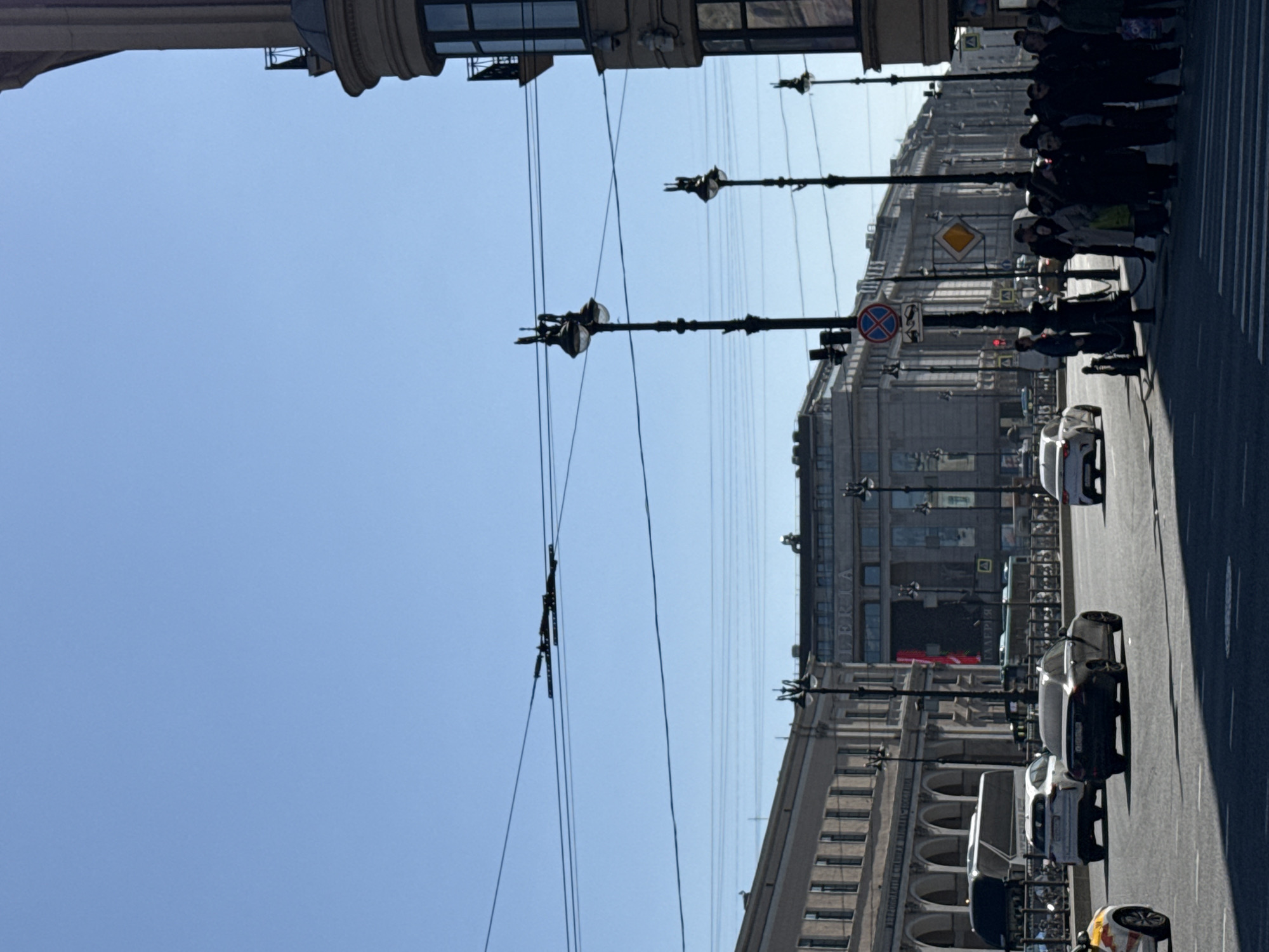The image depicts a street scene in Saint Petersburg, Russia. The view is oriented vertically, looking along Nevsky Prospect. Buildings with classical architecture line the street, including the "Lekka Galleria" building on the right. Power lines crisscross the sky. Streetlights extend from the buildings. Pedestrians are gathered at a crosswalk, waiting to cross. Several cars are parked or moving along the street. The sky is clear and blue. Traffic signs are visible, including one that prohibits a certain action.
