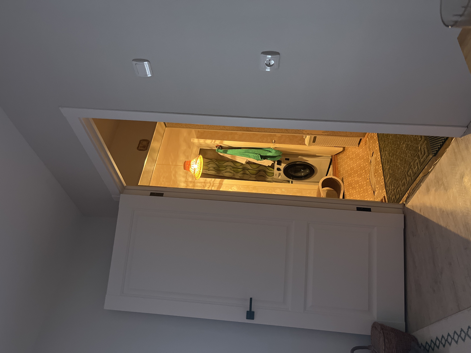 The image captures a high-angle view looking down into a small utility or laundry area, seen from above a partially open white door. The perspective suggests the camera is positioned in a hallway or room, looking into a slightly recessed or smaller adjacent space.
Inside the utility area, a white front-loading washing machine is visible on the right side. Above the machine, a green towel hangs on a rod. A lamp with an orange or yellow woven shade hangs from the ceiling, casting a warm, bright light over the space. The walls of this interior section are covered with a patterned wallpaper, featuring floral or geometric designs in earthy tones. The floor exhibits a mix of textures, including what appears to be a patterned mat or rug.
In the foreground, above the doorway, two ceiling-mounted electrical outlets are visible, consistent with European-style sockets. The room from which the photograph is taken has plain white walls and ceiling. The white door, featuring recessed panels and a dark-colored handle, is a central element, framing the view into the utility space.
The lighting indicates an indoor setting, likely reliant on artificial illumination, possibly in the evening. There are no people visible, and no specific activity is taking place. The scene depicts a functional, organized household area, providing a glimpse into a residential interior, consistent with a home in Chaikovsky, Russia. No visible text is present.