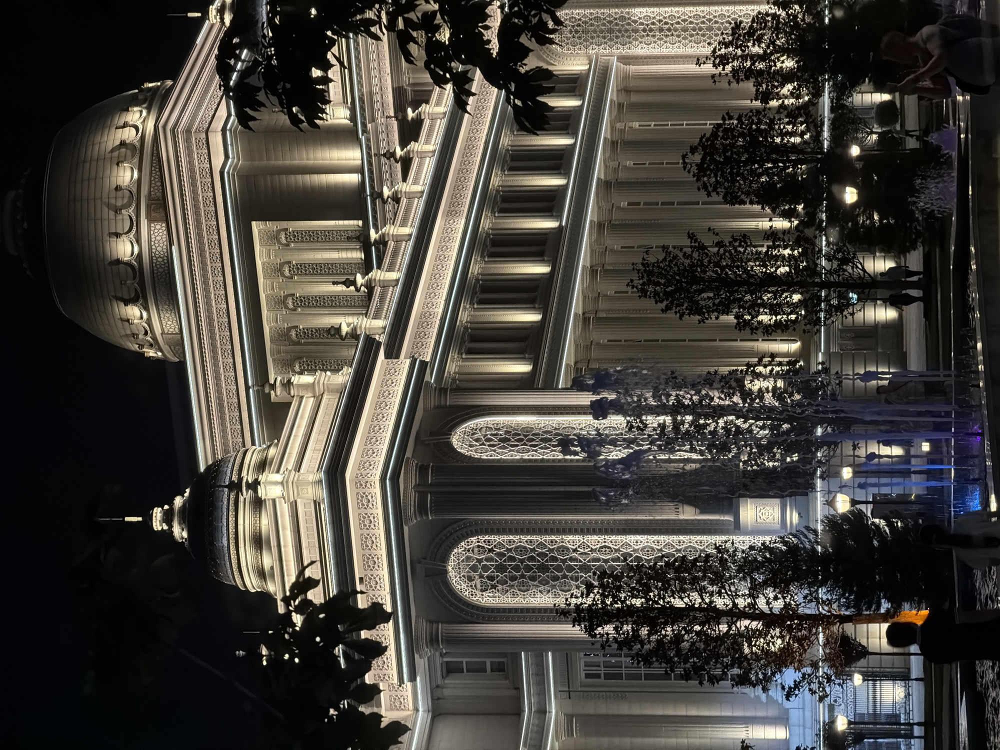The image depicts the National Museum of Tajikistan in Dushanbe, Tajikistan at night. The imposing building is illuminated by warm, white lights, highlighting its classical architecture with large domes, columns, and ornate window designs. In the foreground, a fountain with colorful, illuminated water jets is visible, with several trees silhouetted against the bright facade. A few people are present in the scene, some standing near the fountain and others walking in the background. The overall atmosphere is one of grandeur and nighttime illumination.