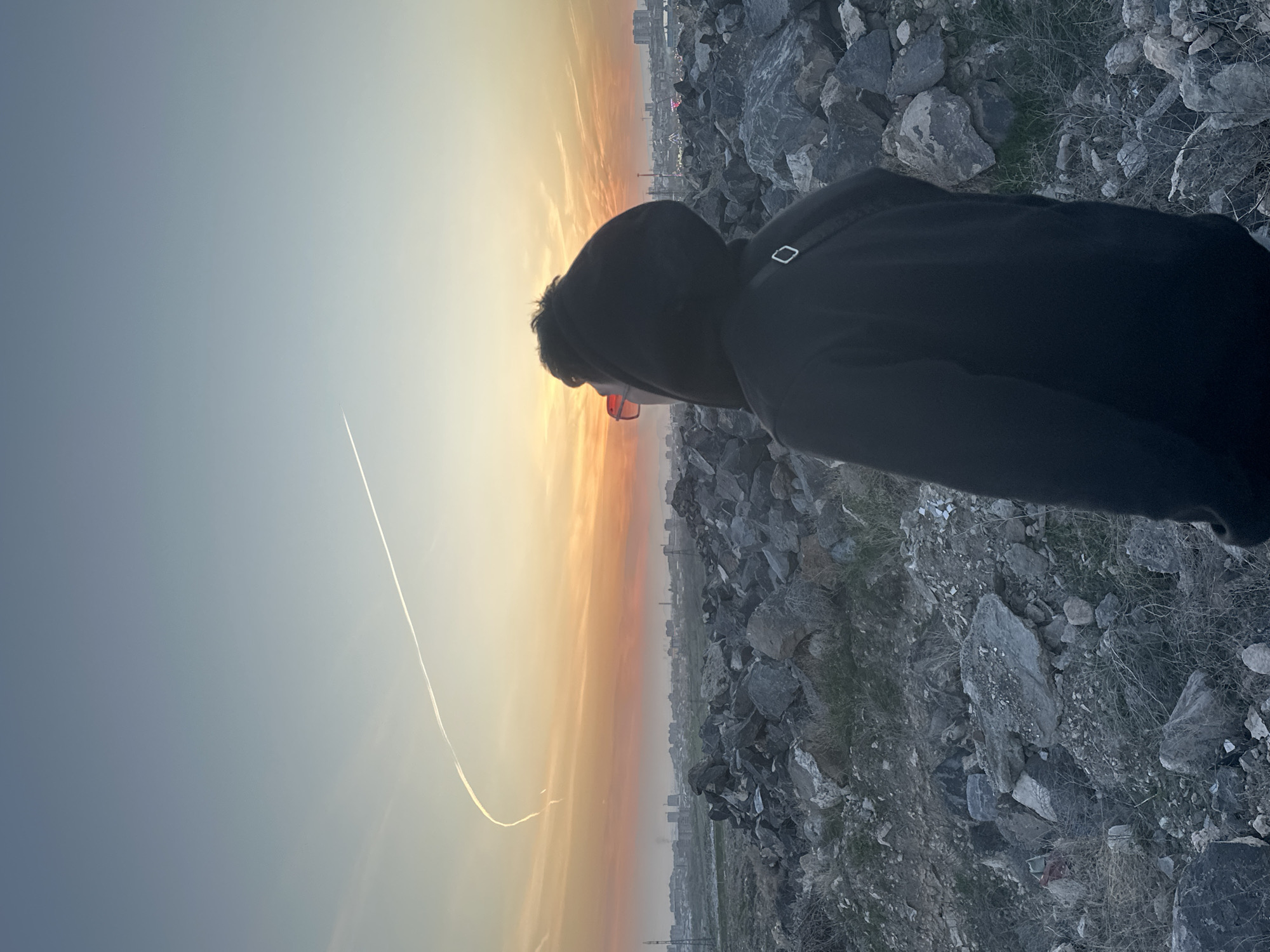 The image depicts a person wearing a black hoodie and red-tinted glasses, standing on a rocky hillside at sunset. The person is facing towards the sunset, which fills the sky with orange and yellow hues. In the distance, a city skyline is visible. A contrail is visible in the sky. The foreground is dominated by rocks and sparse vegetation. The person appears to be observing the sunset and the city in the distance.