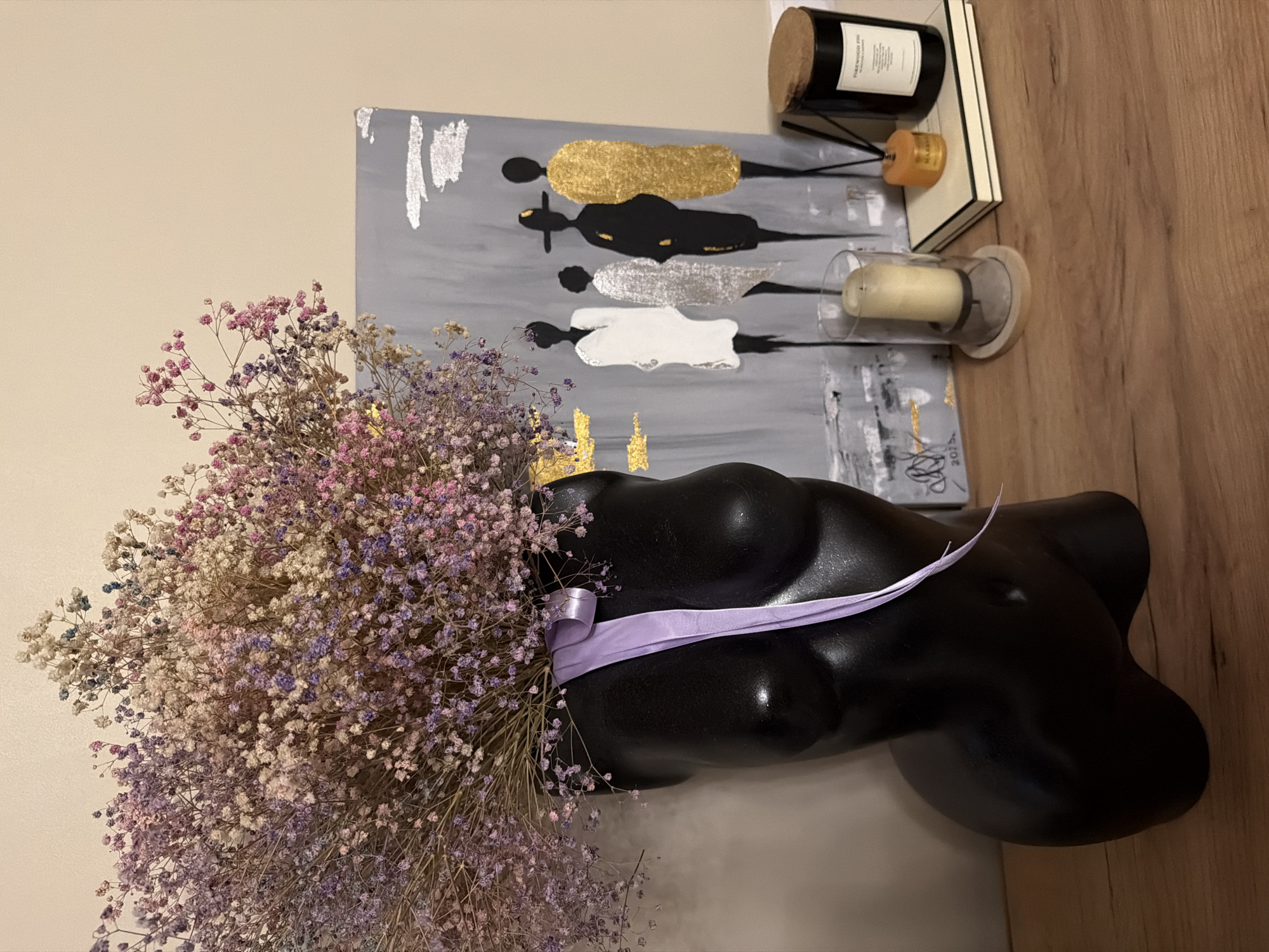 The image shows a still life arrangement with a decorative black torso sculpture in the foreground, adorned with a lilac ribbon. A large bouquet of dried baby's breath flowers, in shades of pink, purple, and white, is placed in front of the sculpture. Behind the sculpture and flowers is a piece of abstract art with a gray background, featuring stylized figures in gold, black, and silver. On a wooden surface to the right of the art, there are several decorative items: a black candle in a jar with a label, a small orange scented candle, and a cylindrical glass holder containing a white candle. The overall scene is indoors, with a plain wall in the background and a wooden surface, suggesting a home or studio setting. There is no discernible time of day or weather information. The style of the art and the objects suggest a modern and artistic aesthetic.
Visible text:
- On the black candle jar: "FIREWOOD PINE" and other unreadable text.
- On the abstract art, near the bottom right corner: "2023."