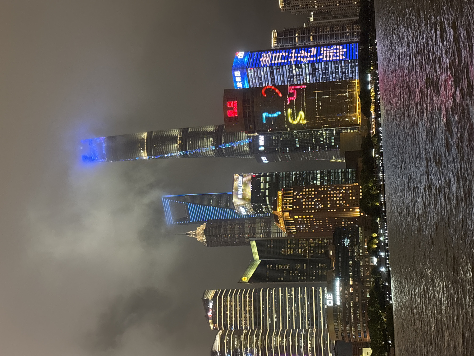 The image depicts the Lujiazui financial district in Shanghai, China, at night. A body of water, likely the Huangpu River, occupies the foreground, reflecting the lights of the city. The skyline is dominated by numerous illuminated skyscrapers, their facades adorned with various colors and patterns. Notably, the Shanghai Tower, the tallest building, is visible with a blue light illuminating its upper section and a vertical streak of blue lights running down its side. Other prominent buildings include the Shanghai World Financial Center with its distinctive trapezoidal opening, and the Jin Mao Tower with its ornate spire. Buildings are adorned with glowing signs and advertisements, contributing to the vibrant urban nighttime scene. Dense fog or clouds partially obscure the tops of some of the taller structures, adding a dramatic atmosphere. Trees and lights line the riverfront, suggesting a promenade or park area. No people are visible in the image.