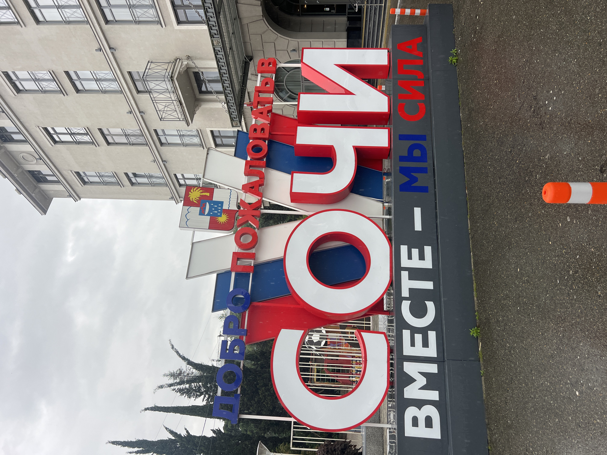 The image depicts a large sign that reads "Добро пожаловать в Сочи – Вместе Мы Сила" (Welcome to Sochi – Together We Are Strong). The letters are large, colorful, and three-dimensional, with a combination of blue, red, and white. The sign is located in Sochi, Russia. The flag of Sochi is visible next to the sign. An urban environment is visible in the background, including a building with windows and a balcony. An orange traffic cone is near the sign. The ground appears to be wet asphalt.
