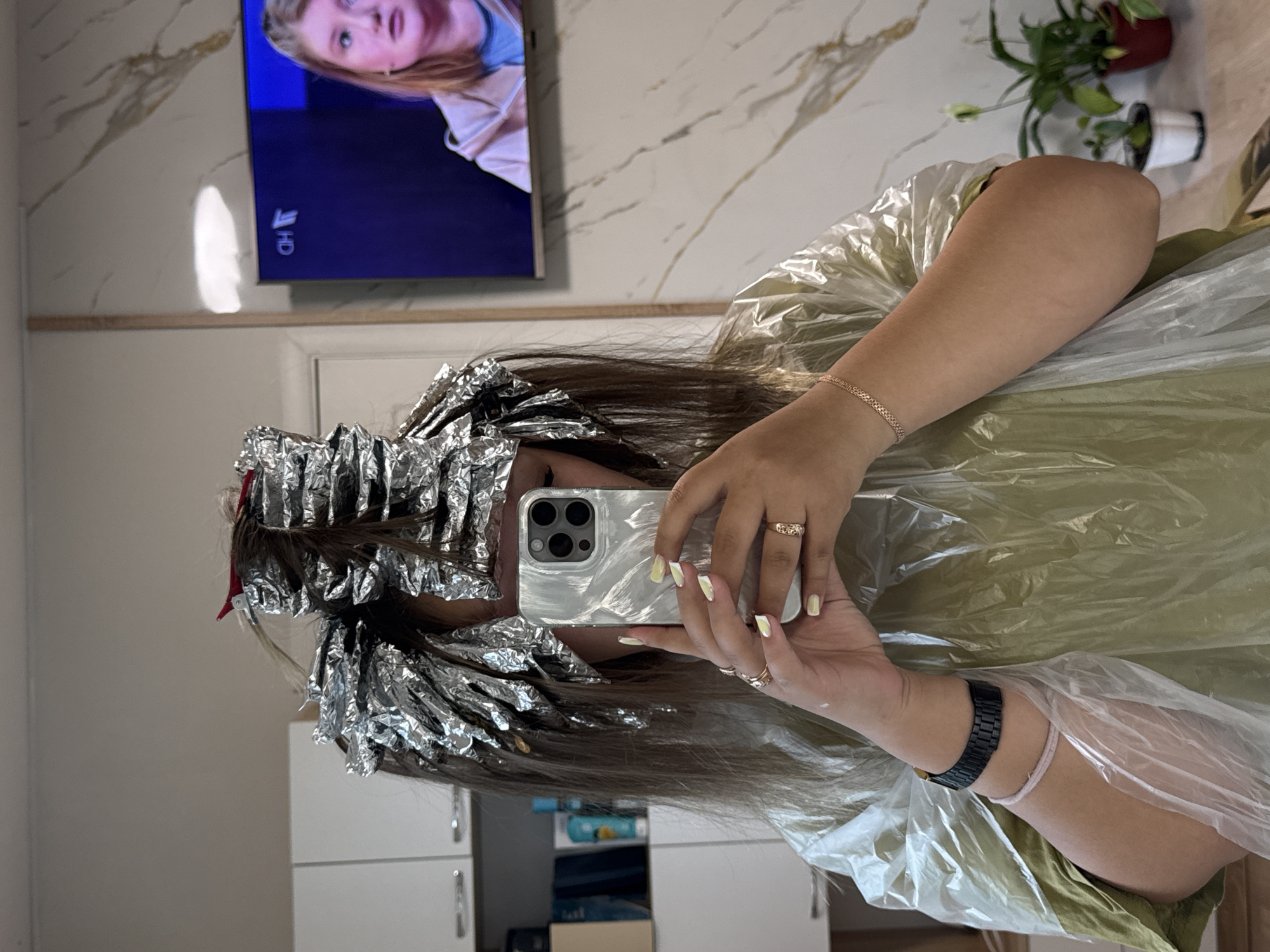 A person is taking a selfie in what appears to be a hair salon. The person is wearing a disposable cape and has their hair wrapped in foils. The person is holding a smartphone, using it as a mirror. The selfie is taken indoors. A television is mounted on the wall, a plant is visible in the background. No information is available in the photo to confirm the city or country.