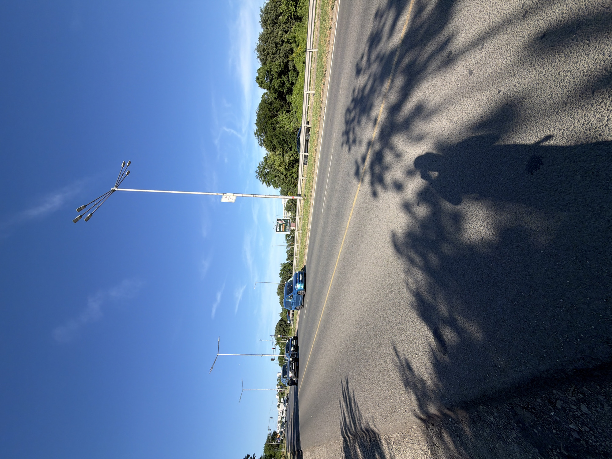 This media file captures a scene from a roadside in Burgas, Bulgaria, under a clear blue sky with sparse wispy clouds. The perspective suggests the photo was taken by someone standing on the side of a paved road, looking towards the road and a line of trees beyond a guardrail. The time of day appears to be late afternoon, indicated by the long, dappled shadows cast by trees onto the asphalt.
The main subjects are the road itself, a series of streetlights, and several cars traveling along the road. The streetlights, with multiple lamps each, are prominently featured against the bright sky. Several cars are visible, including a bright blue vehicle in the foreground and a line of darker cars further down the road. Beyond the road and guardrail, a dense line of green trees forms a backdrop. In the distance, more streetlights and what appear to be roadside advertisements or signs can be seen.
The activity taking place is simply the movement of traffic along a road. The weather is sunny and pleasant. There are no clear emotional cues, and the scene is one of everyday travel.
Notable details include the distinct shadows of tree leaves and branches creating a natural pattern on the road. The presence of multiple streetlights suggests this is likely a highway or a main thoroughfare. A partially visible sign with text in what appears to be Cyrillic script can be observed near the blue car, adding a specific location cue. The overall impression is a bright, clear day on a road in a populated area, likely near a city or town.