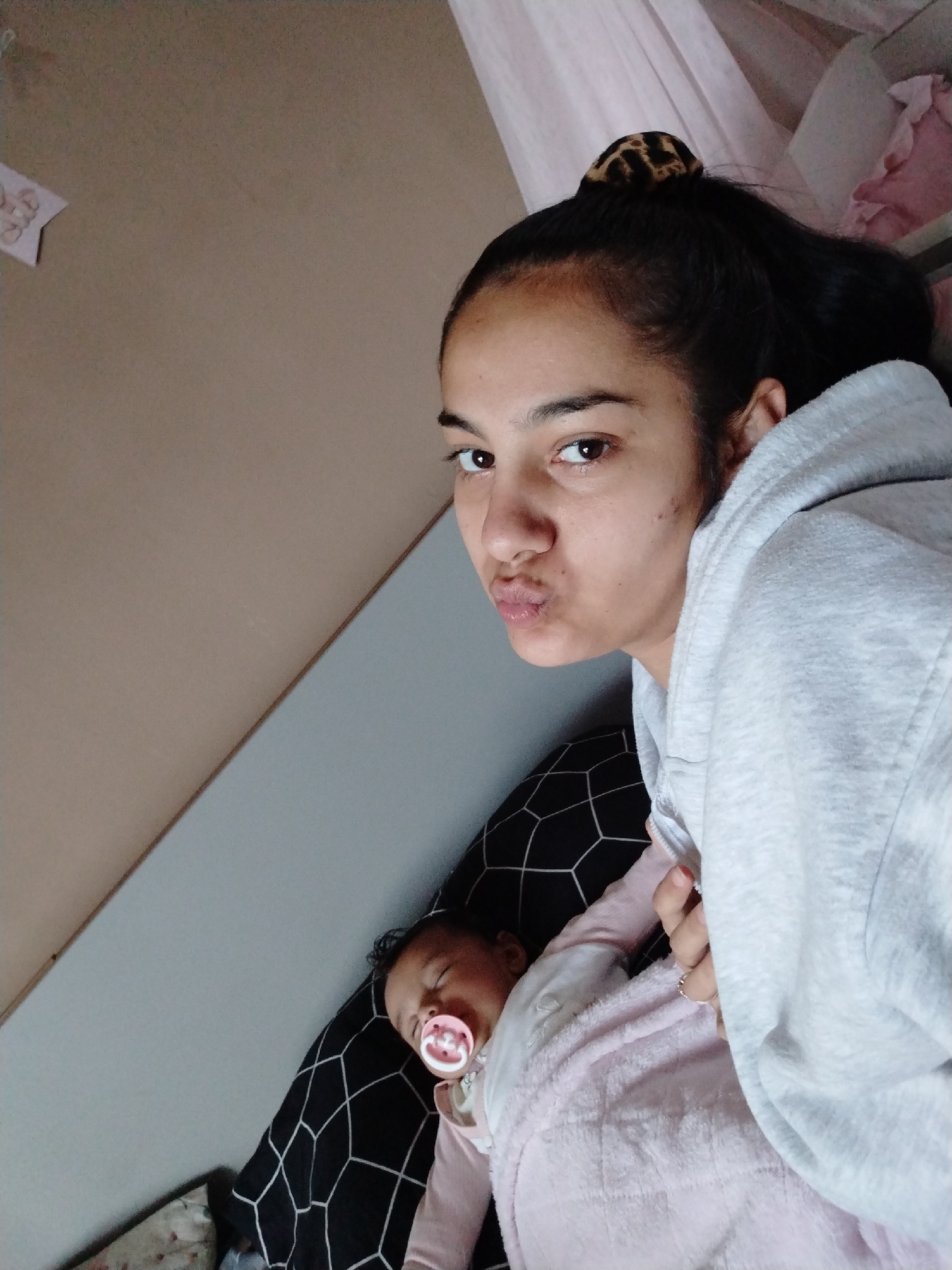 A person with dark hair pulled into a high ponytail, secured with a leopard print scrunchie, is positioned on the right side of the image, puckering their lips. They are wearing a light gray hooded sweatshirt. Next to them, a baby is sleeping on a black and white patterned pillow with a pink pacifier in its mouth, wrapped in a pink blanket. The background consists of a two-toned wall, with the top half in a warm beige and the lower half in a cooler gray. The location is Unknown, Unknown.
