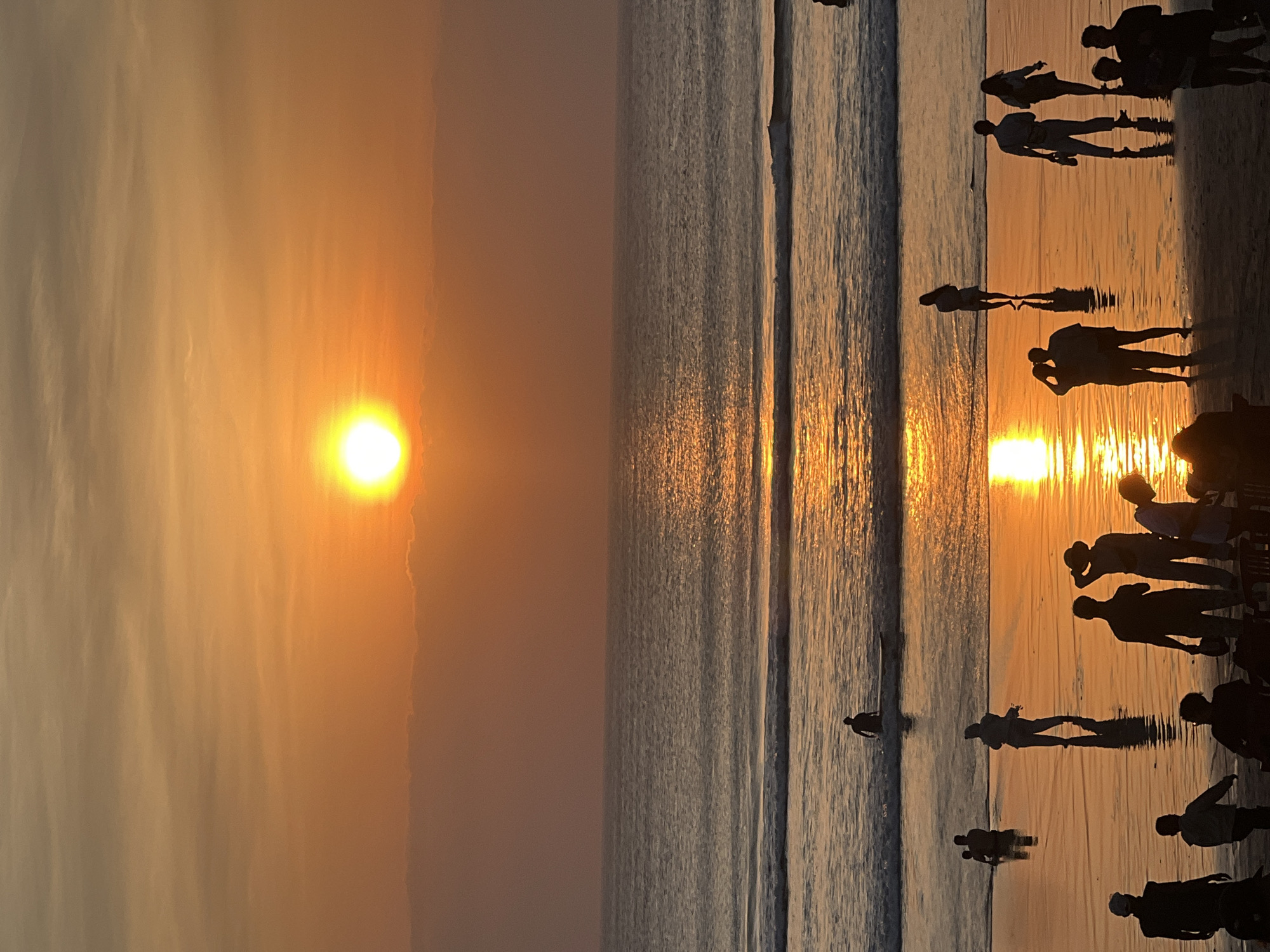 This image depicts a sunset scene at Kuta Beach, Bali, Indonesia. The sun, a bright orb, is setting over the horizon, casting a warm, orange glow across the sky and reflecting on the water. Gentle waves are rolling onto the shore, creating a shimmering effect on the wet sand. Silhouettes of people are scattered across the beach and in the shallow water, some walking, some standing, and others interacting. A person is visible on a surfboard in the water. The scene captures a serene and picturesque moment of a beach sunset, a common sight in the popular tourist destination of Kuta.
