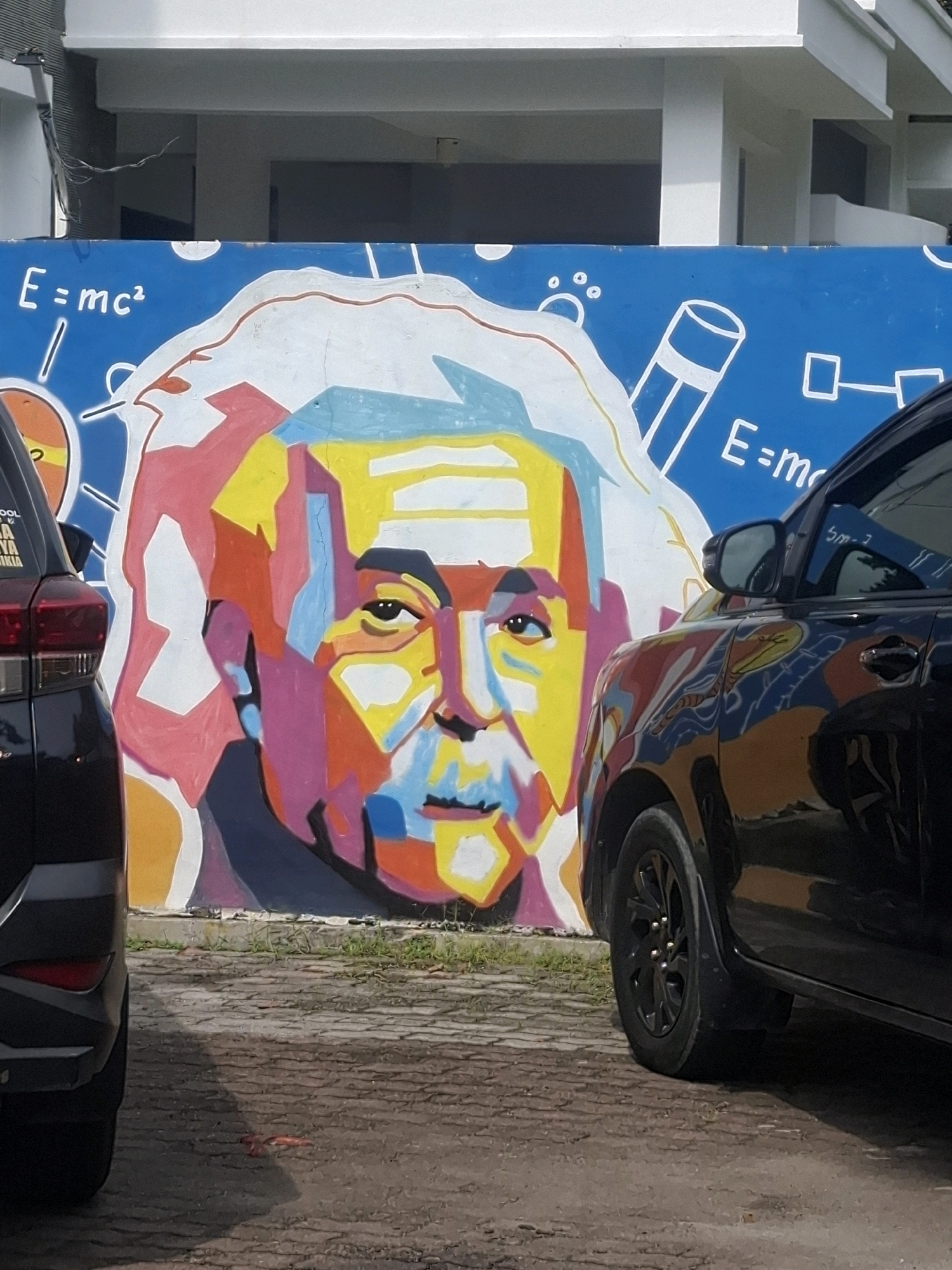 The image shows a mural of Albert Einstein on a blue wall at Medan Estate, Indonesia. The mural is rendered in a cubist style with vibrant colors. Einstein's face is depicted with his characteristic wild hair and mustache. The mural also features scientific formulas, including "E=mc²".
In the foreground, two black cars are parked in front of the mural. The car on the left is partially visible, showing its rear taillight and part of its side. The car on the right is more fully visible, with its side reflecting the colors of the mural. The ground in front of the mural is paved with rough, light-colored stones. Some patches of grass are visible between the stones. The background shows a white building with architectural features such as columns and overhangs. The sky is not visible in the image. The overall impression is that of an outdoor scene, likely in a public space or a parking lot, decorated with artistic murals.