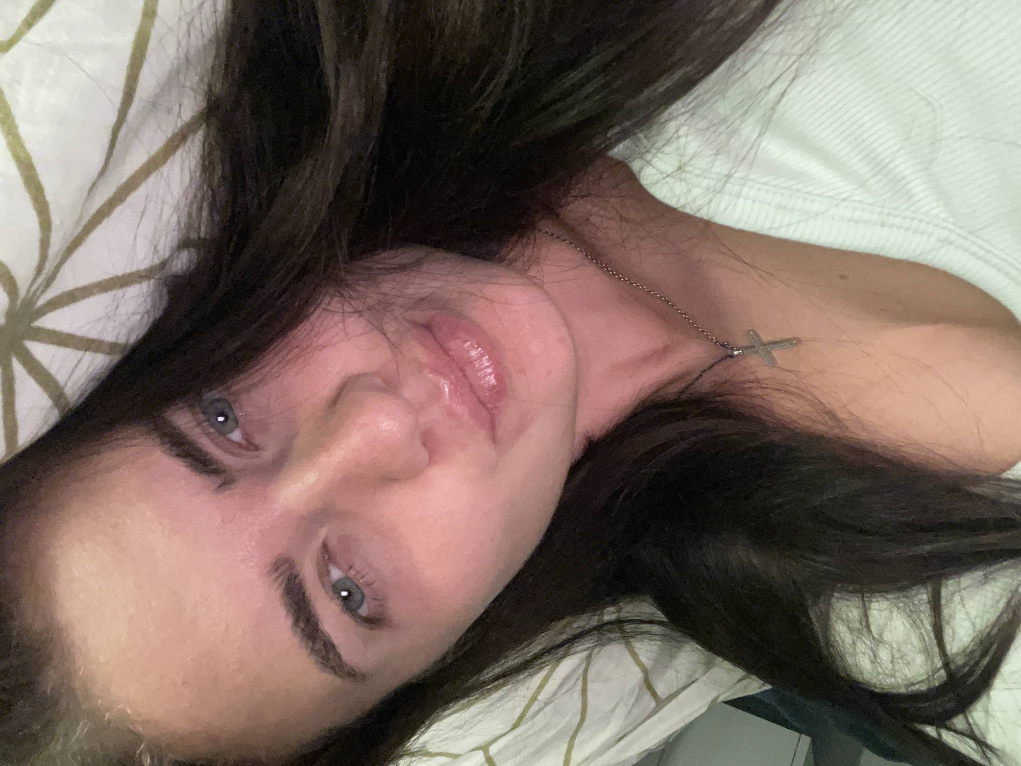 The image is a close-up, overhead shot of a young woman lying down, possibly in a bed, with her dark hair spread around her face and shoulders. She is wearing a light-colored, ribbed top and a silver necklace with a cross pendant. Her eyes are open, and her lips are slightly parted, giving the impression she is about to speak or has just finished. The background appears to be a textured white surface with a pattern, possibly a duvet or pillowcase. There is no indication of the location being in Мосметаллоконструкция Онт, Russia. The lighting suggests an indoor setting, possibly at night or in low light conditions, with a warm, pinkish hue cast over her skin.