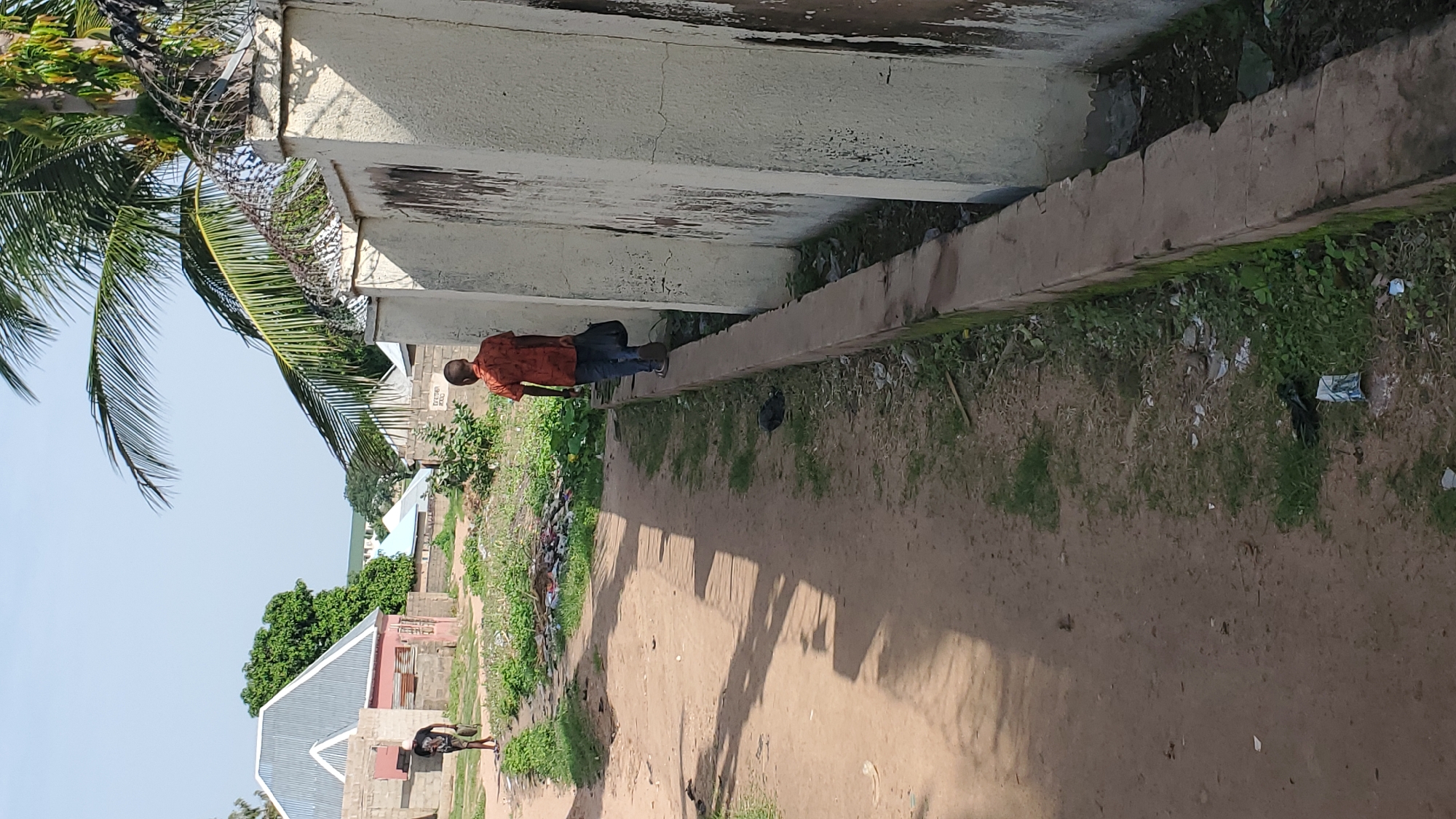 The image depicts a street scene in Jalingo, Nigeria, with two individuals. In the foreground, a young person wearing an orange patterned shirt and blue jeans walks away from the camera, carrying a black bag. They are walking along a raised concrete curb that runs parallel to a textured, light-colored wall topped with barbed wire. The wall appears to be part of a property boundary. To the left of the person, a dirt path is visible, with scattered debris and green vegetation. Further down the path, another person, wearing a dark t-shirt and shorts, is walking towards the left side of the frame. In the background, there are buildings with corrugated metal roofs and cinder block construction, surrounded by trees. The sky is clear and blue. The overall environment appears to be a developing urban or semi-urban area.