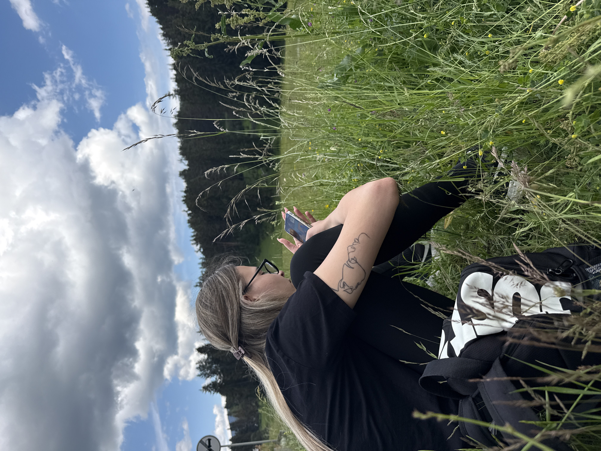 A young woman with long blonde hair tied back with a clip and wearing glasses, a black t-shirt, and black leggings is sitting in tall grass, looking at her phone. She has a tattoo on her left arm. A black backpack with white lettering is beside her. The background shows a dense forest of dark green evergreen trees under a partly cloudy blue sky. The location is in the city of Yundola, Bulgaria.