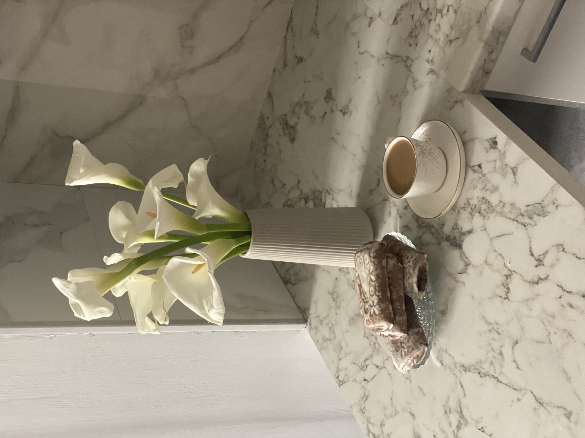 A still life composition is presented on a marbled countertop. In the foreground, a small glass dish holds a stack of four rectangular pastries decorated with white icing patterns. To the right of the pastries, a white ceramic cup with a gold rim sits on a matching saucer filled with a light brown beverage, likely coffee. Behind the pastries and coffee, a tall, ribbed, light gray vase contains a bouquet of white calla lilies with green stems. The background features a marbled wall and a portion of a white cabinet. The scene is illuminated by ambient light, casting soft shadows. The location is not specified within the image.