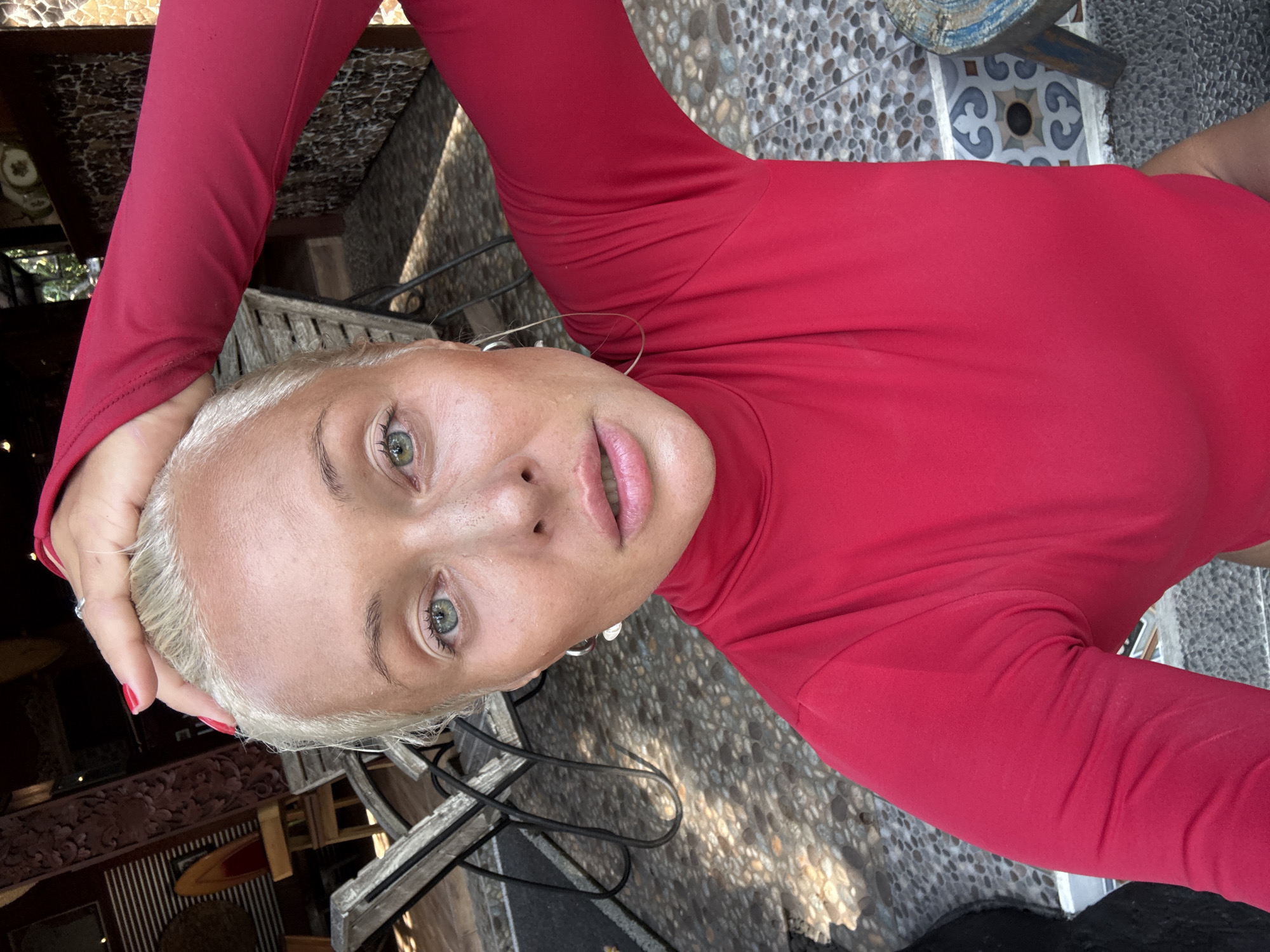 A fair-skinned woman with blonde hair and green eyes is facing the camera. She is wearing a red long-sleeved shirt. Her right hand is resting on her head. The background includes patterned flooring, a wooden structure with carvings, and some foliage. The location and specific city are not identifiable from the image.