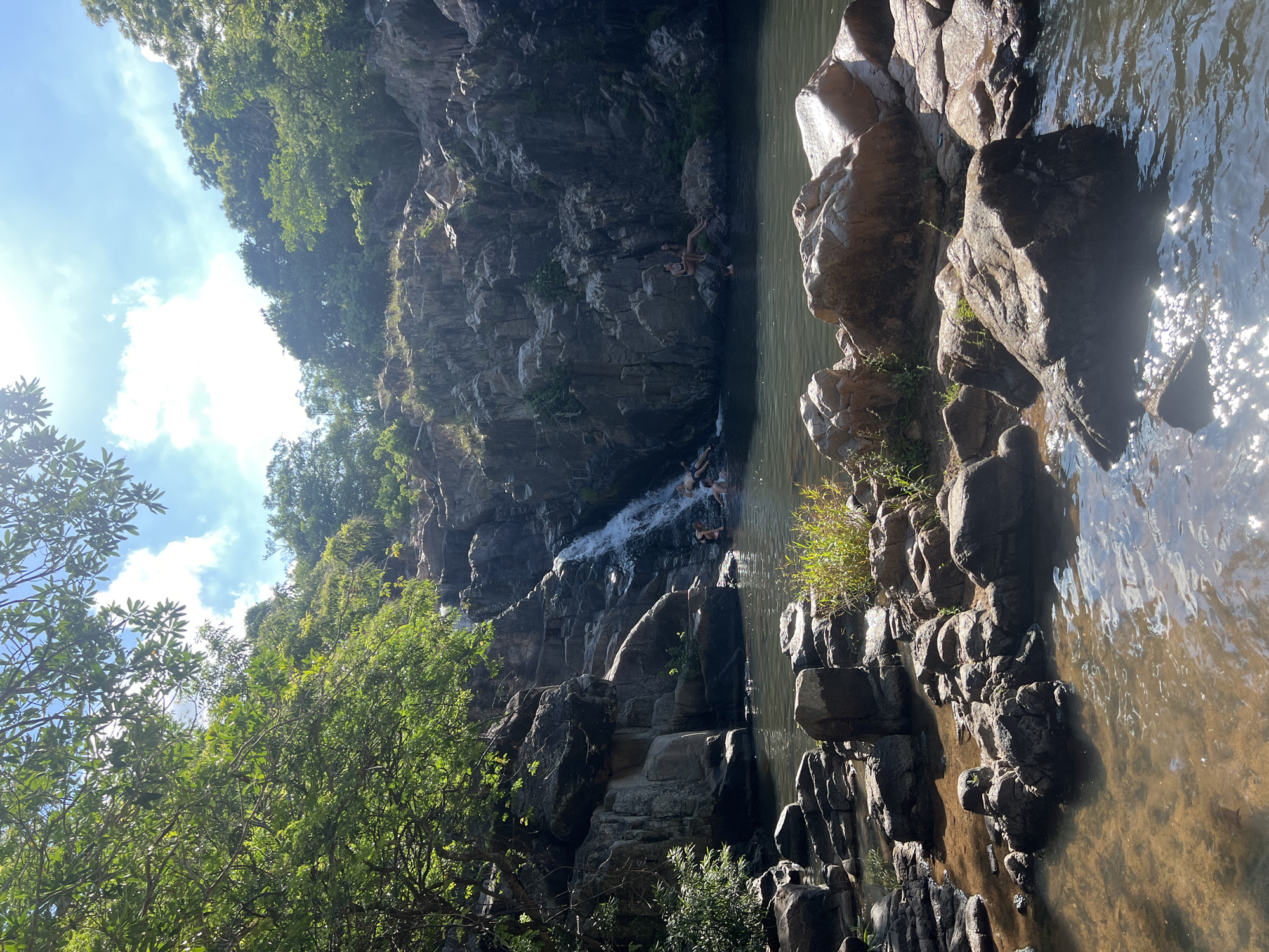 A serene natural scene unfolds with a waterfall cascading down a rocky cliff face into a clear, dark pool of water. Lush green trees and foliage frame the top and sides of the image, with sunlight dappling through the leaves and reflecting on the water's surface. Several individuals are present in and around the water, appearing to be enjoying the refreshing environment. Two people are seated on rocks to the right of the waterfall, while others are visible in the water near the falls, possibly swimming or wading. The rocky terrain extends into the foreground, with various sized rocks scattered along the water's edge. The overall impression is one of a tranquil, natural oasis, potentially a popular spot for recreation and relaxation. The location is identified as Vĩnh Hải, Vietnam.
