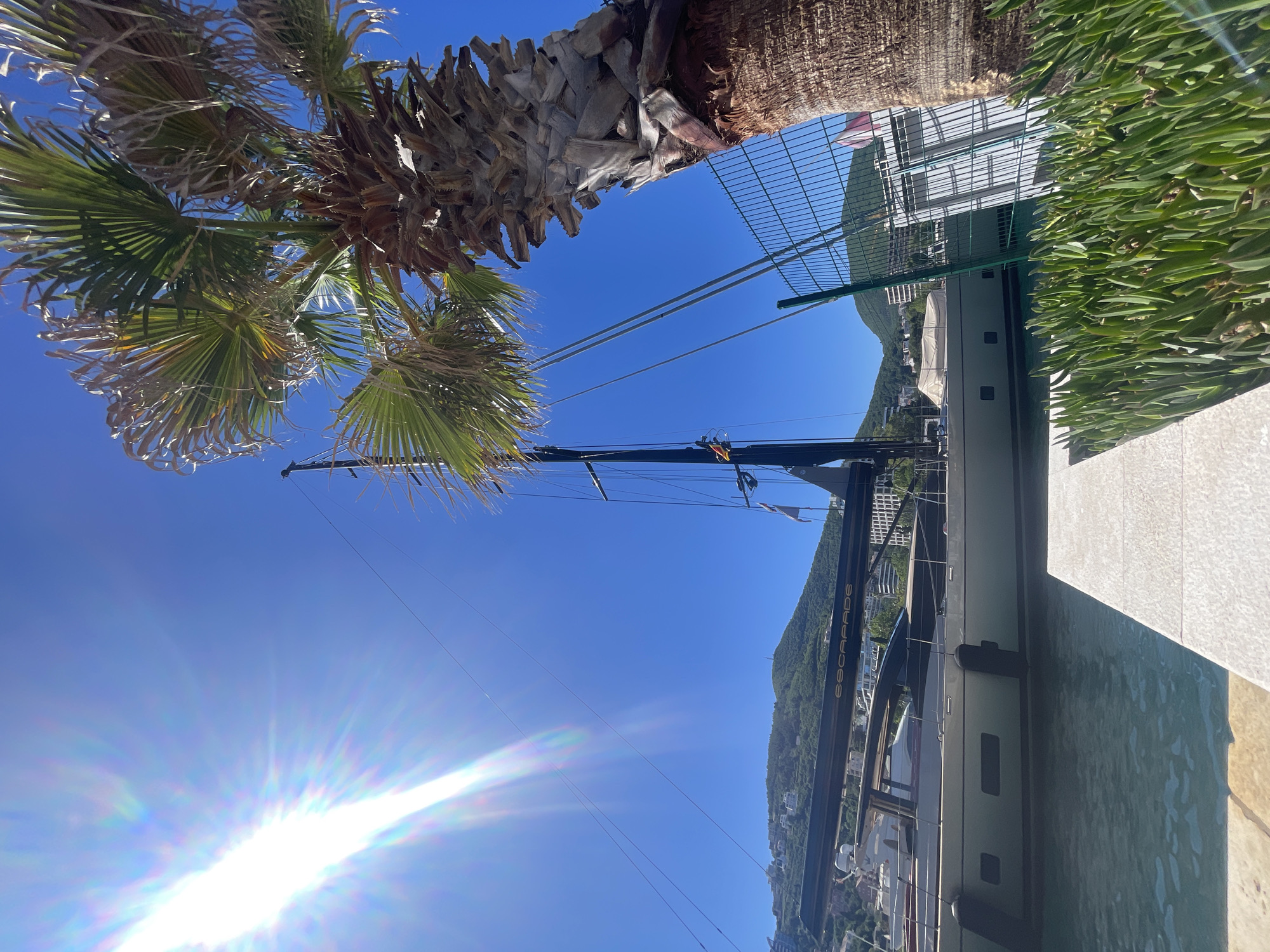 The image depicts a daytime scene featuring a large yacht named "ESCAPADE" docked at a marina, with a lush, green hillside in the background. The sky is a clear, bright blue with the sun prominently visible in the lower left corner. A palm tree is visible in the upper left, while greenery lines the upper right of the frame. The yacht is positioned adjacent to a concrete quay. A wire fence is behind the yacht. The location appears to be a coastal area, with a cityscape nestled in the hillside. The specific city and country are not identifiable from the image.