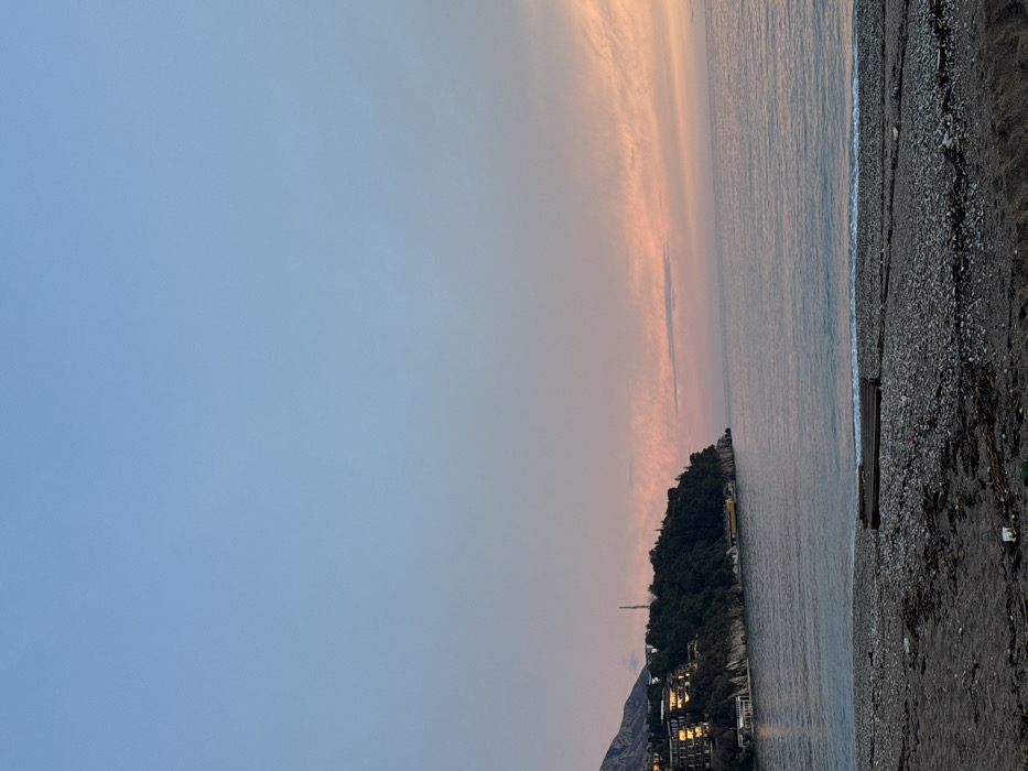 This image captures a serene coastal landscape at dusk, set in Budva, Montenegro. The sky dominates the upper portion, transitioning from a clear light blue on the left to soft pink and orange hues towards the right, indicative of a sunset or sunrise. Subtle, wispy clouds are visible within the warmer sky tones.
A calm sea occupies the middle and right sections, gently reflecting the sky's colors. Small waves ripple along the shoreline. On the left, a dark, forested peninsula or hill extends into the water, covered densely with green trees. At its base, several buildings with illuminated windows glow softly, suggesting hotels or residences. A slender tower rises from the higher ground of the landmass, and a larger mountain range is visible in the distant background.
To the far right, a dark, pebbly or gravel beach stretches along the coast, with some scattered debris. A wooden bench or platform is positioned on the beach near the water. The scene is devoid of people, creating an atmosphere of tranquility and natural beauty. The clear skies and still water suggest calm weather conditions. No visible text is present in the image.