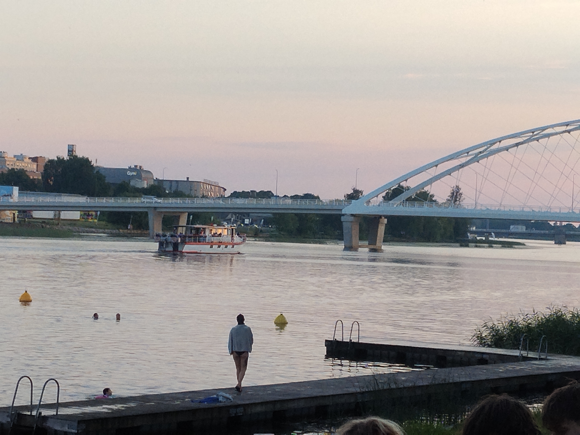 The image captures a tranquil scene at dusk in Pärnu, Estonia. In the foreground, a person in a light-colored jacket and dark swimwear stands on a concrete pier, facing away from the camera and towards the water. To the left of the pier, there are metal ladder rungs leading into the water. In the middle ground, several individuals are partially submerged in the calm river, with only their heads visible. A yellow buoy floats in the water to the left of the pier. Further out in the river, a white passenger boat with an orange stripe is cruising, its upper deck populated with passengers. In the background, a distinctive white arched bridge spans the river, with a multi-lane road visible on top. The cityscape of Pärnu is visible on the distant left bank, featuring various buildings, including one with the word "Gym!" prominently displayed. The sky above is a soft gradient of pink and light blue, indicating either sunrise or sunset. The overall atmosphere is serene and depicts a leisurely evening by the water in Pärnu.