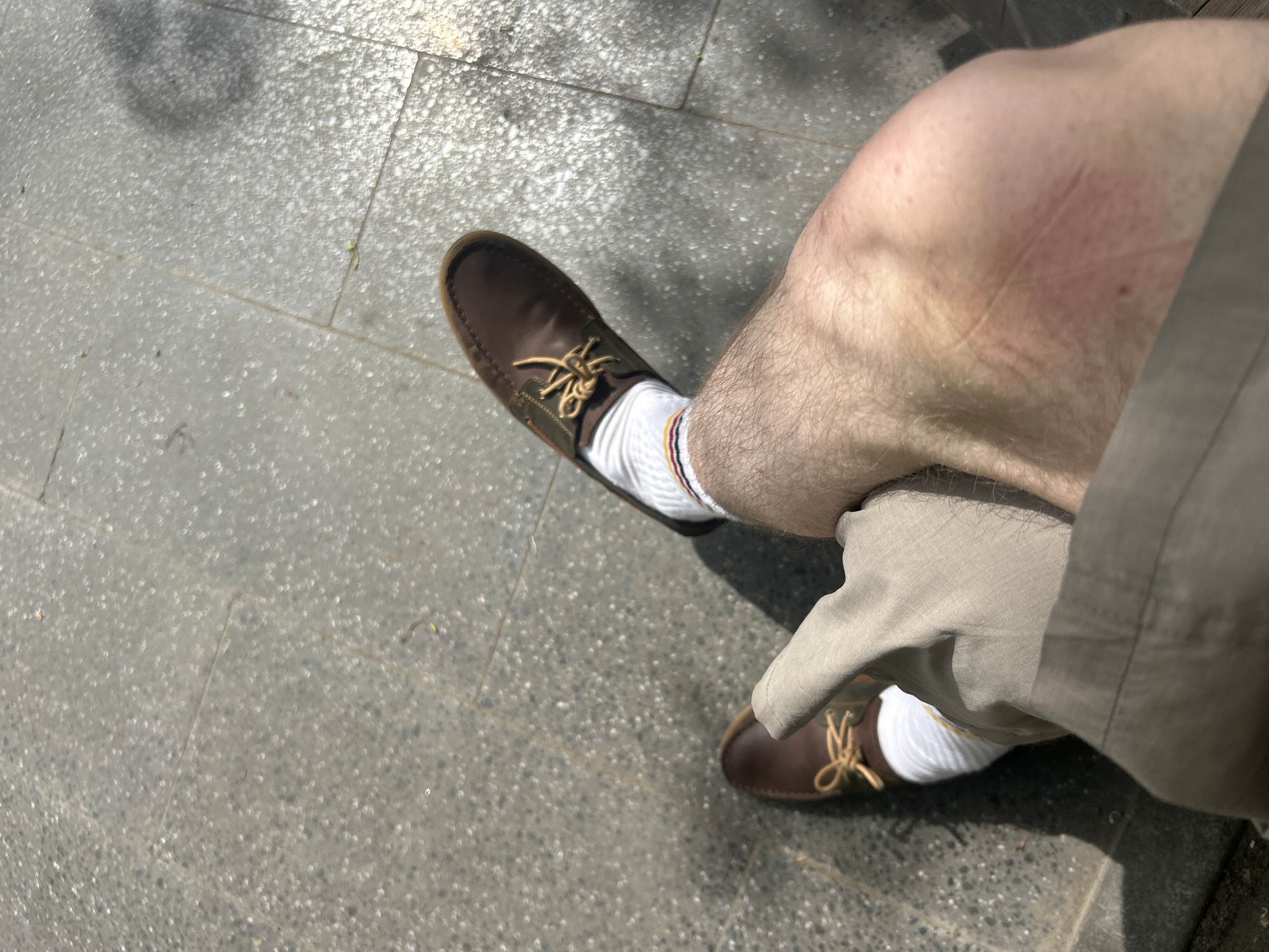 A first-person perspective shot shows a person sitting outdoors. The lower half of the person's body is visible, including a bare leg with a reddish patch on the knee, khaki shorts, white socks with red and gold stripes at the top, and brown leather shoes with untied laces. The person is sitting on a gray concrete surface, possibly a sidewalk or patio. The location is in an unknown city.