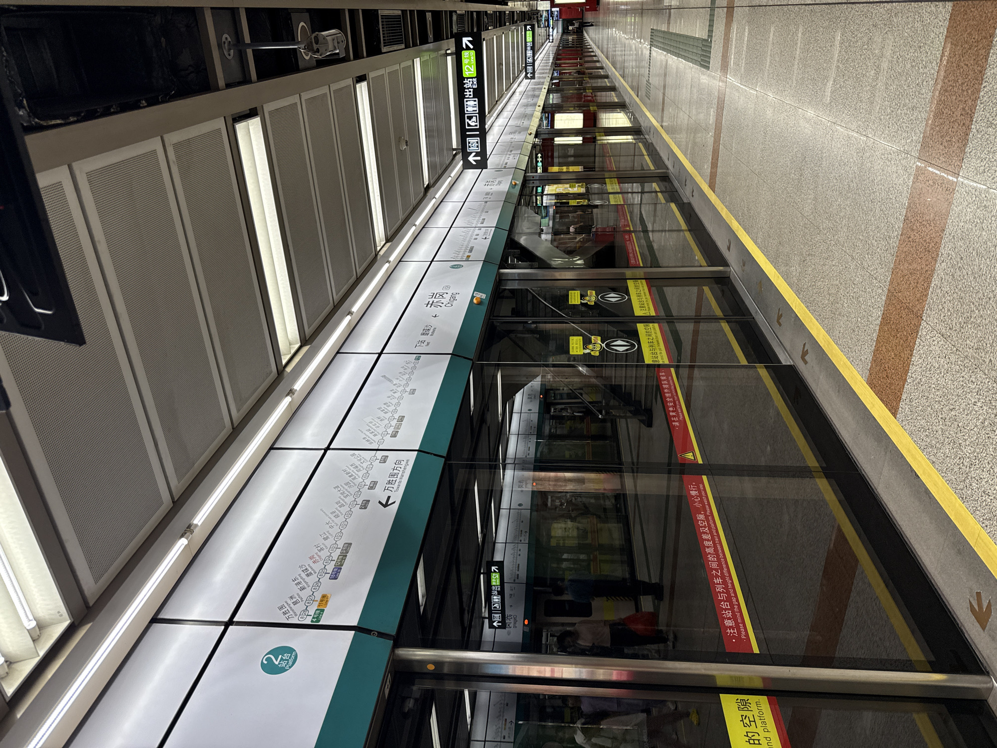 The image depicts the interior of a Guangzhou Metro station in Haizhu District, China. The scene is characterized by a clean and modern design, with a focus on functionality. The platform is well-lit by fluorescent lights mounted on the ceiling. Several train cars are visible, stopped at the platform, with their doors closed. The doors are made of glass, reflecting the station's environment.
On the platform, there are signs indicating train routes and station names. One prominent sign displays "2号线 站台" (Line 2 Platform) along with a list of stations. Another sign shows "万胜围" (Wanshengwei) with an arrow pointing to the right, indicating the direction of travel. There are also signs with icons for accessibility features like elevators and restrooms, as well as directional arrows for exits.
Yellow safety lines are marked on the platform floor, near the edge of the platform, to warn passengers of the train's proximity. Red and brown striped patterns are embedded in the floor tiling, adding a subtle visual element. Several people are present on the platform, some waiting for trains, while others are disembarking or walking. One individual is visible in the background, walking along the platform.
The train cars themselves have a sleek, metallic exterior with green accents along the top. The windows of the train cars are tinted. A warning sign with a triangular exclamation mark and text in Chinese and English reads "请注意站台与列车之间的高度差及空隙,小心慢行。" (Please mind the gap and height difference between train and platform. Please watch your step.) This indicates a common safety precaution in subway systems. The overall impression is of an efficient and organized public transportation hub.