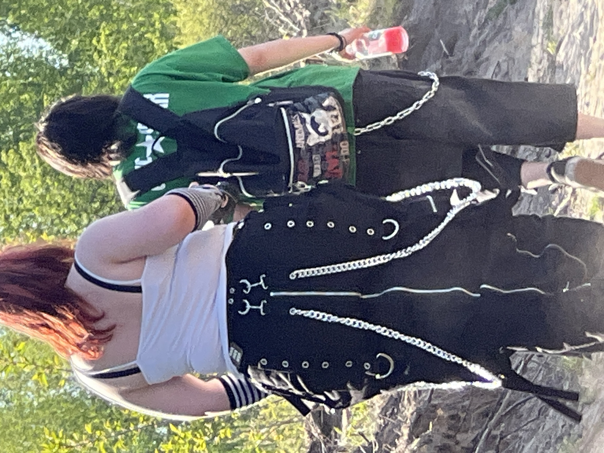 The image shows two people standing outdoors. One person, wearing a green shirt, black shorts with a chain, and a backpack, is holding a water bottle. The other person is wearing a white tank top and a black garment with chain and silver hardware detailing. They appear to be standing in a natural, possibly wooded area. The location is presumed to be in Ciemne, Poland.