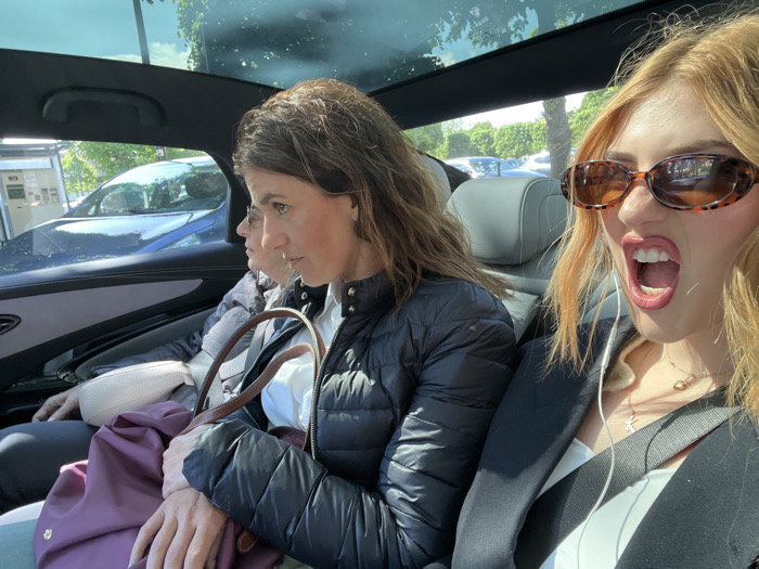 The image shows three women inside a car. The woman in the back seat is wearing a white shirt, black jacket, and sunglasses. She has red hair and is looking at the camera, with her mouth open in a loud yell. She is wearing a thin, white wire in her ear, which is likely an earbud. The middle woman is wearing a black jacket and a purple handbag. She is looking at something off-camera, and her left hand is holding the handbag. The woman in the front seat is partially visible. She is wearing a white shirt and a black jacket. She is looking at the middle woman, and her left hand is resting on her lap. The car is a dark color, and there is a view of the street outside through the windows. The location is Vernon, France, with the coordinates 49.091 latitude and 1.485 longitude. The city is known for its charming streets and historical sites, and the image suggests a casual, urban setting. The scene is likely an everyday occurrence, with the women engaged in a conversation.