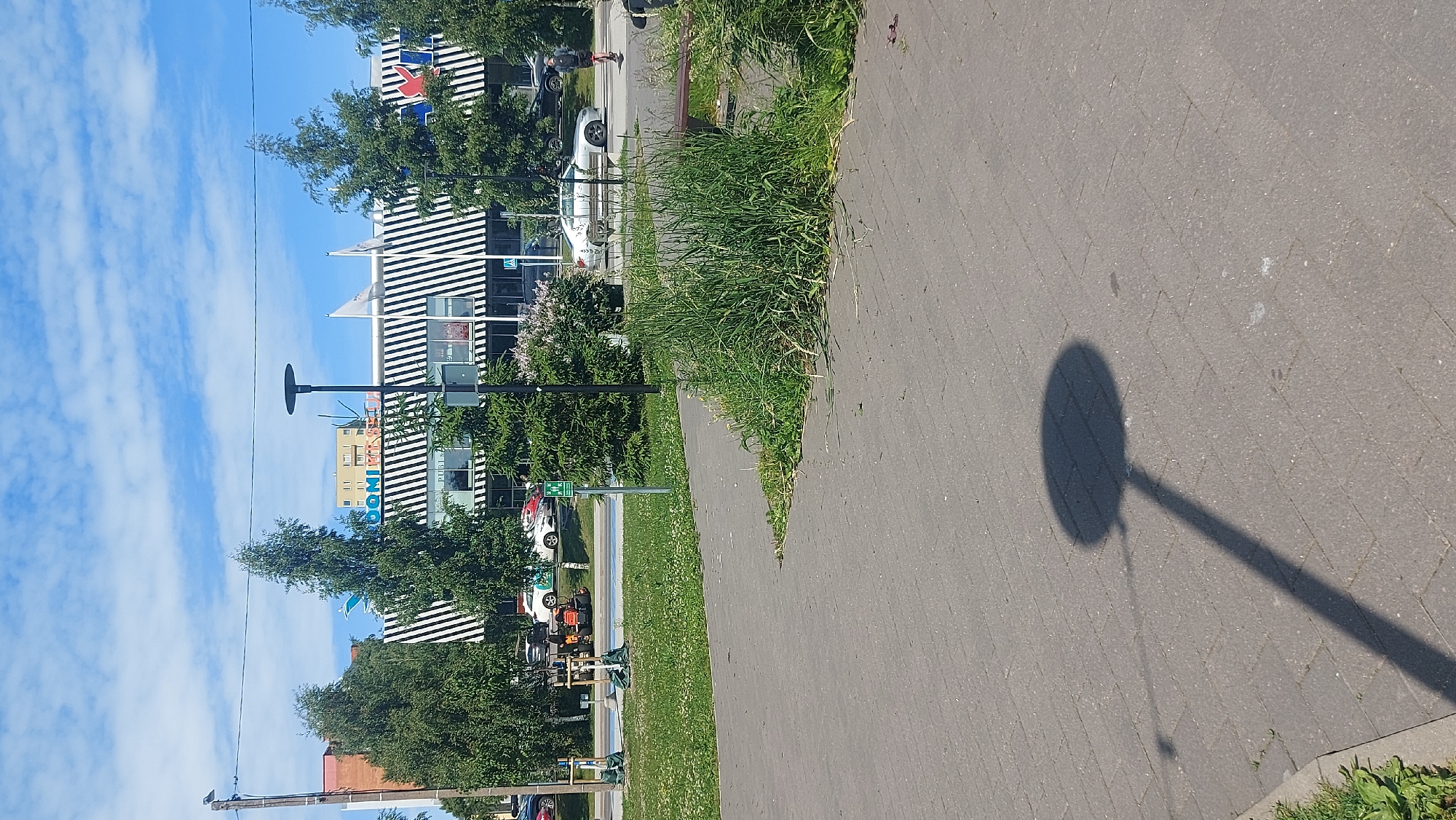 The image captures a street scene in Tallinn, Estonia, on a bright, partly cloudy day. In the background, a modern building with a distinctive black and white striped facade stands prominently. Above the building, a yellow, multi-story structure is visible, suggesting a commercial or residential area. Several trees, some tall and leafy, others smaller evergreens, dot the landscape in front of the building.
In the foreground, a paved walkway made of gray interlocking bricks stretches across the frame. To the right of the walkway, a patch of overgrown green grass with some flowering weeds spills out, partially obscuring a wooden bench. The shadow of a streetlamp, with a circular top and a long pole, is cast diagonally across the paved surface, indicating the direction of the sun.
On the walkway, a person on a red electric scooter is moving away from the camera towards the right. Another person is walking in the distance on the right side of the image. Cars are visible on a road behind the trees and in front of the building, including a white sedan, a silver sedan, and other vehicles. A small orange tractor-like vehicle is parked on the grass to the left, next to some trees that appear to be recently planted and secured with stakes and netting. A green pedestrian sign with a wheelchair symbol is also visible near the center of the image. The overall impression is a well-maintained urban environment with signs of recent landscaping or construction.