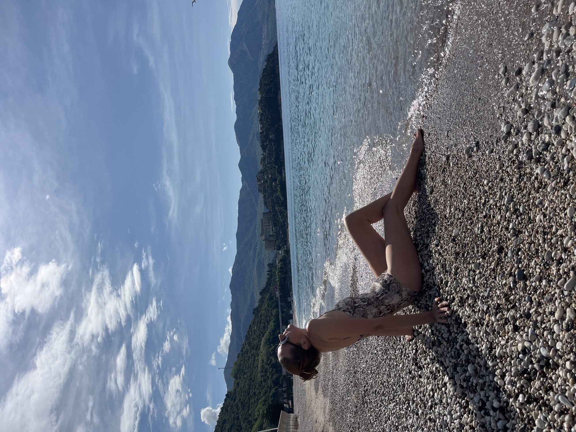 The image displays a woman relaxing on a pebbled beach at the water's edge, under a bright blue sky. She is lying on her back, propped up on her elbows, with her head tilted towards the sky, wearing a patterned one-piece swimsuit and sunglasses on her head. Her legs are partially visible, bent and crossed.
The foreground is dominated by the pebbled beach, where countless small, smooth, multi-colored stones meet the clear, blue-green water. Gentle waves lap at the shore, catching the sunlight and creating shimmering reflections. In the mid-ground, the expansive body of water stretches towards a lush green coastline.
The background features towering, verdant mountains that rise directly from the shore, extending into the distance. Some buildings are visible nestled among the trees at the base of the mountains, suggesting a coastal town or resort. Above, the sky is a vibrant blue with scattered white clouds, some wispy and some more defined. A small bird is faintly visible flying high in the sky.
The scene depicts a sunny day, likely morning or late afternoon, with strong natural light illuminating the landscape. The overall mood is one of tranquility and natural beauty, consistent with a vacation or leisure setting in a coastal area like Budva, Montenegro. There is no visible text in the image.