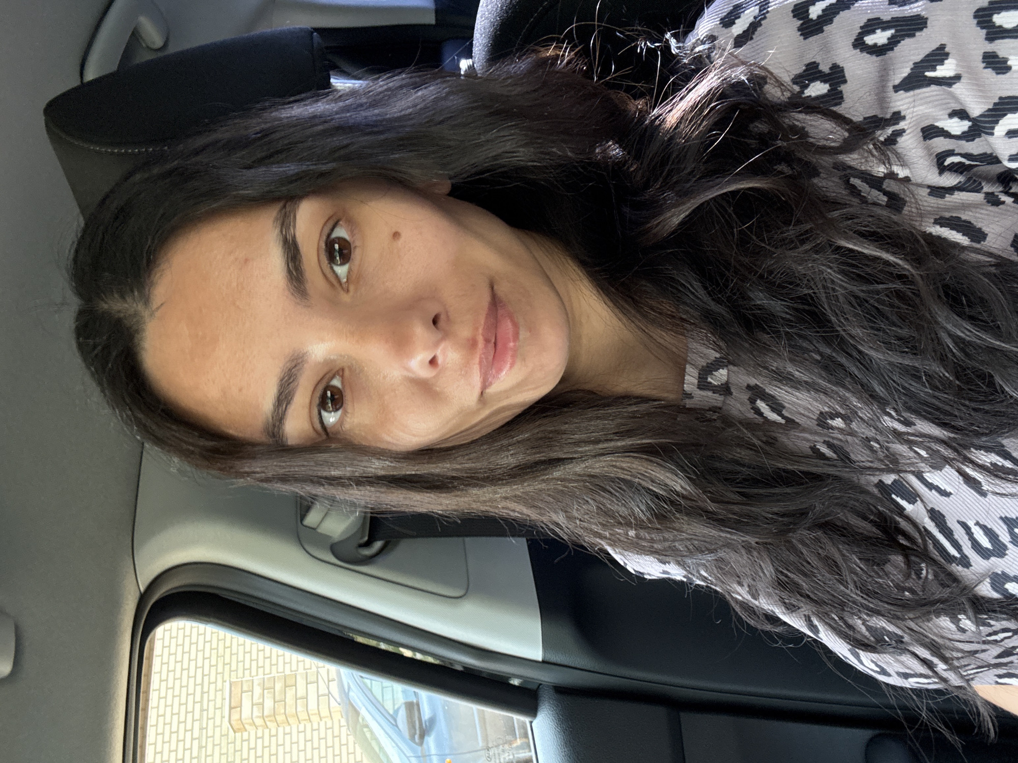 A woman with long, dark, wavy hair is sitting in the passenger seat of a car, facing the camera. She is wearing a light-colored top with a black and white leopard print. Her skin is fair, and she has brown eyes. The interior of the car is visible, including a black headrest and a gray door panel. Outside the car window, a brick building and a blue car can be seen. The image appears to have been taken during the daytime, with sunlight casting shadows on the woman's hair.