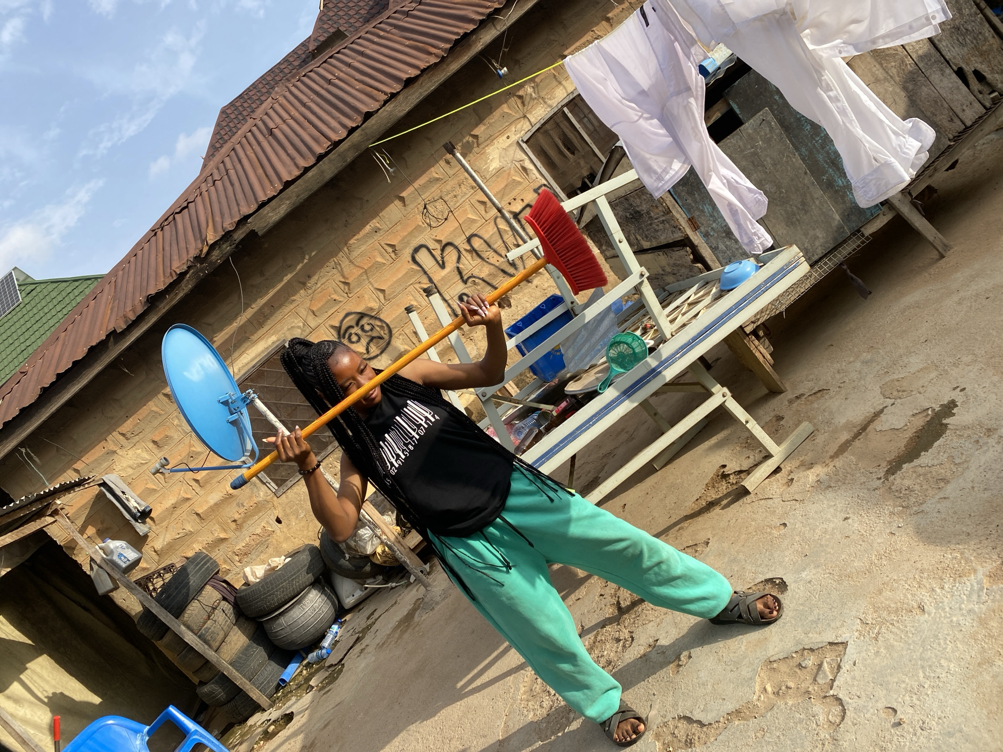 A young woman with braided hair, wearing a black tank top and teal pants, is sweeping a concrete area with a red-bristled broom. She is outdoors, with a brick building and a corrugated iron roof in the background. A blue satellite dish is mounted on the building. In the foreground, white clothing is hanging on a clothesline. The scene appears to be in Jos, Nigeria, a city known for its temperate climate and rocky landscape. The overall impression is one of domestic activity in a somewhat utilitarian setting.