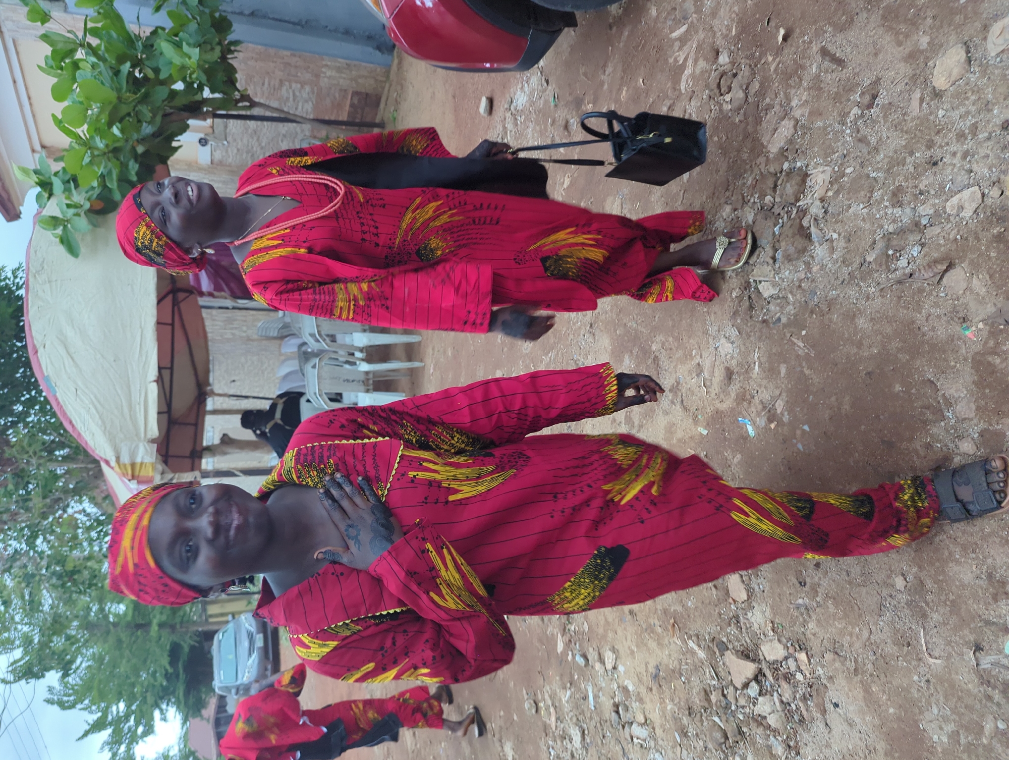 Two women, possibly young adults, are prominently featured in an outdoor setting in Unguwar Rimi, Nigeria, captured in a portrait-style photograph. They are both dressed in vibrant, matching traditional attire, consisting of long garments made of red fabric adorned with striking yellow and black patterns, featuring both linear stripes and abstract, feather-like designs. Each woman also wears a headwrap/turban made from the same patterned fabric.
The woman on the left, closer to the foreground, is smiling broadly towards the camera, with her left hand placed gently on her chest. Her skin is dark, and an intricate, dark henna or tattoo design is visible on her left hand and wrist. She wears dark sandals.
The woman on the right stands slightly behind and to the side of the first, also smiling and looking towards the camera. She carries a black handbag over her left shoulder and wears golden-colored flip-flops. Her outfit's neckline is visible, showing a simple necklace.
The background suggests a casual, semi-urban environment. The ground is unpaved, covered in dirt and scattered small rocks. Parts of a red vehicle (possibly a car or motorcycle) are visible behind the woman on the right, along with some green foliage from trees or bushes. In the left background, a light-colored tent or awning and white plastic chairs can be seen, hinting at a gathering or event. No specific time of day is clearly indicated, but the lighting appears natural and soft, suggesting daytime. There is no visible text in the image.