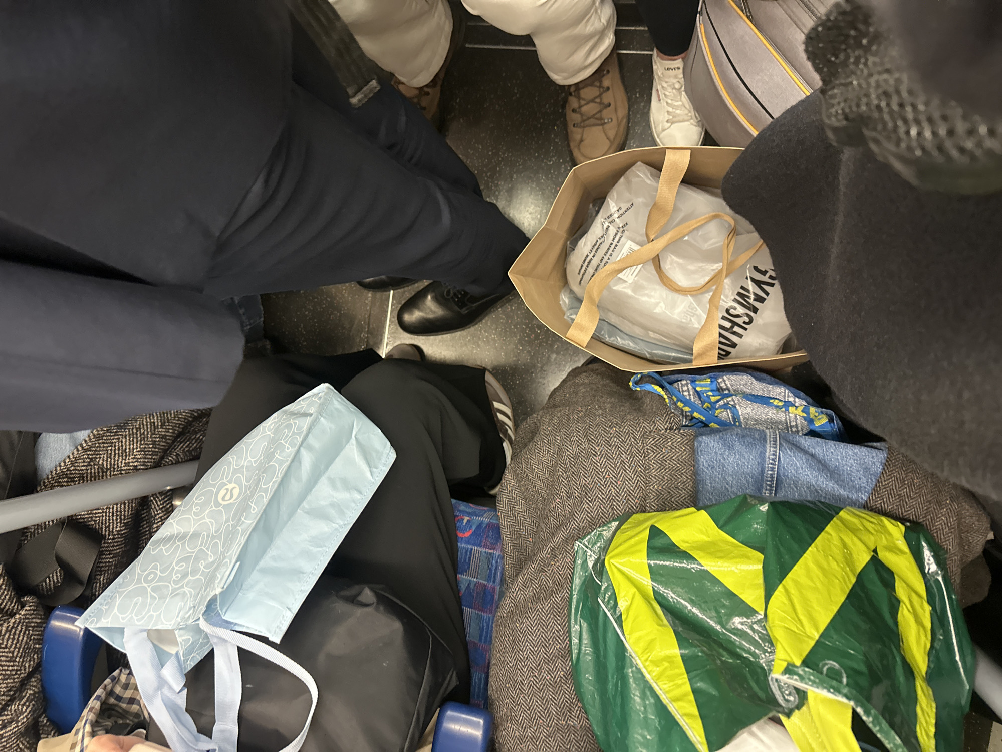 The image depicts the interior of a crowded public transport carriage, consistent with a London Underground train, within the city of London, United Kingdom. The scene captures the lower torsos, legs, feet, and personal belongings of multiple individuals from a downward perspective.
Visible individuals include one person wearing dark trousers and black leather lace-up shoes, standing center-right. To their right, another wears light-colored trousers and brown lace-up boots, while a third exhibits white sneakers with a partially visible "LEVI'S" logo. On the far left, black trousers and Adidas brand sneakers are discernible. A lap covered in dark fabric at the top-left holds a light blue Lululemon shopping bag adorned with white abstract patterns.
Various objects occupy the space. A large, crinkled plastic bag featuring prominent green and yellow diagonal stripes rests on a brown herringbone-patterned textile in the lower-left, adjacent to a folded piece of blue denim. A tan paper shopping bag sits on the dark gray floor in the lower-middle, containing a plastic-wrapped item bearing an inverted "GYMSHARK" logo and warning text. A blue and yellow patterned woven bag and a gray soft-sided bag with yellow and black stripes are partially visible in the lower-right. A vertical metallic pole is present on the left side. The dark gray floor and sections of a blue and red patterned seat fabric are also discernible beneath items.