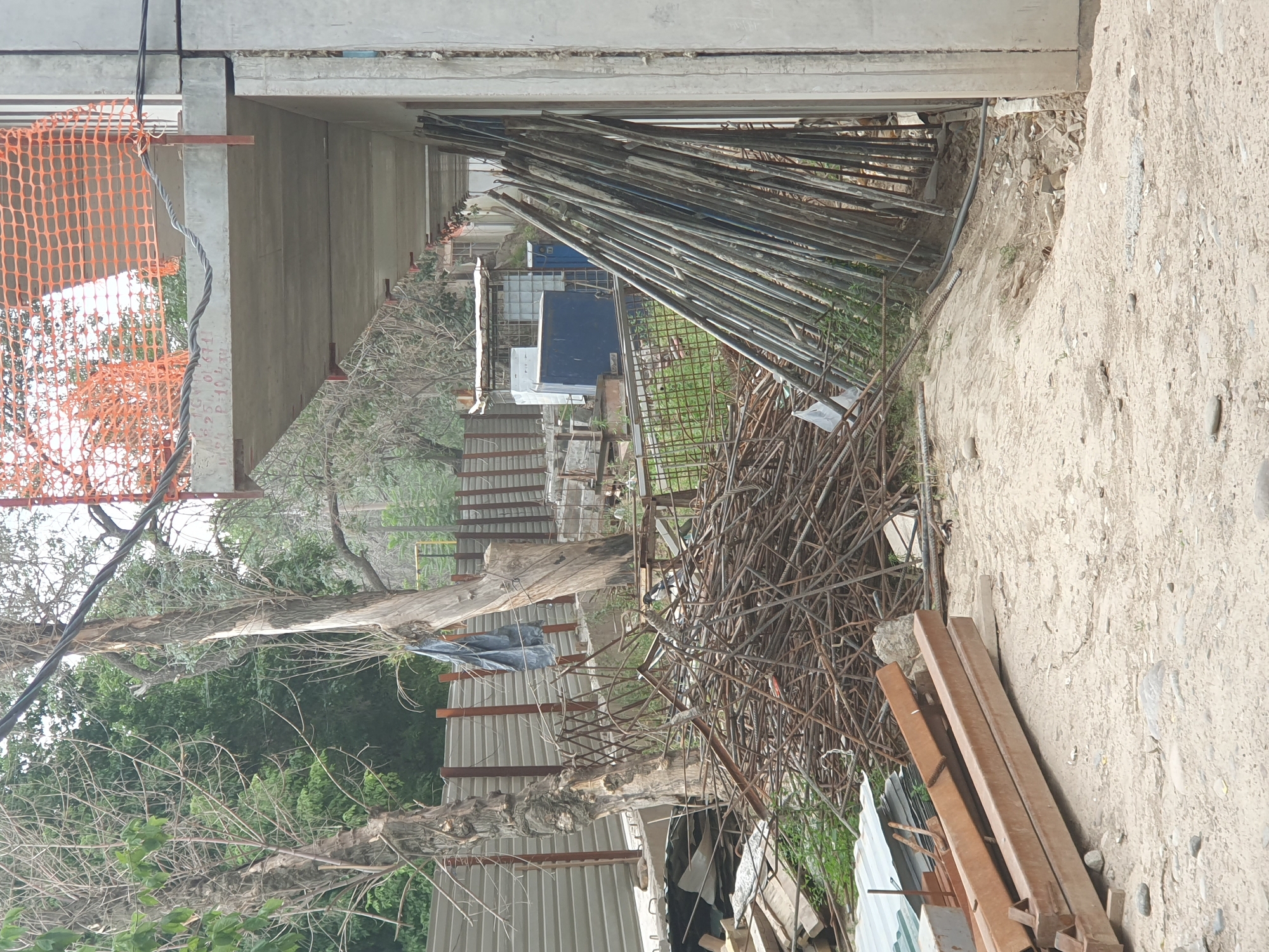 The image depicts a construction site in Tashkent, Uzbekistan. In the foreground, a pile of rusted metal rebar is present. To the right, a stack of long metal poles leans against a concrete building under construction. Behind the rebar, a corrugated metal fence can be seen. Trees with dry branches and some green foliage are visible in the background, along with other construction materials and structures. The ground is covered with sand and small rocks. An orange safety net is strung across the upper portion of the building.