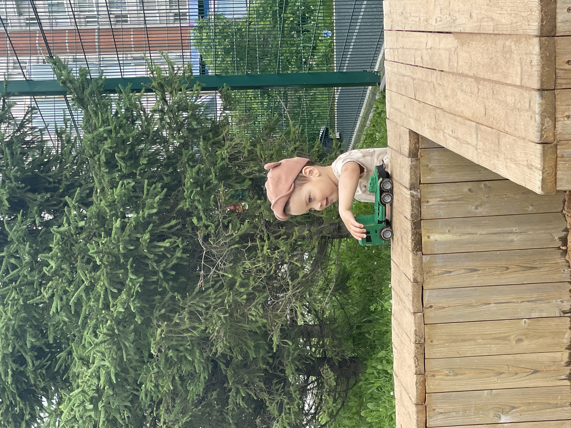 The image shows a young child playing with a toy truck on a wooden structure. The child is wearing a light-colored t-shirt and a light brown cap. They are reaching out to push the green toy truck. The wooden structure appears to be part of a playground or an outdoor play area, constructed from large wooden beams. In the background, there are dense green evergreen trees and a wire fence. Beyond the fence, there are some buildings with windows and what looks like a paved area. The scene is outdoors, and the lighting suggests it is daytime, likely sunny. The child appears engaged and focused on their play. There is no visible text in the image.