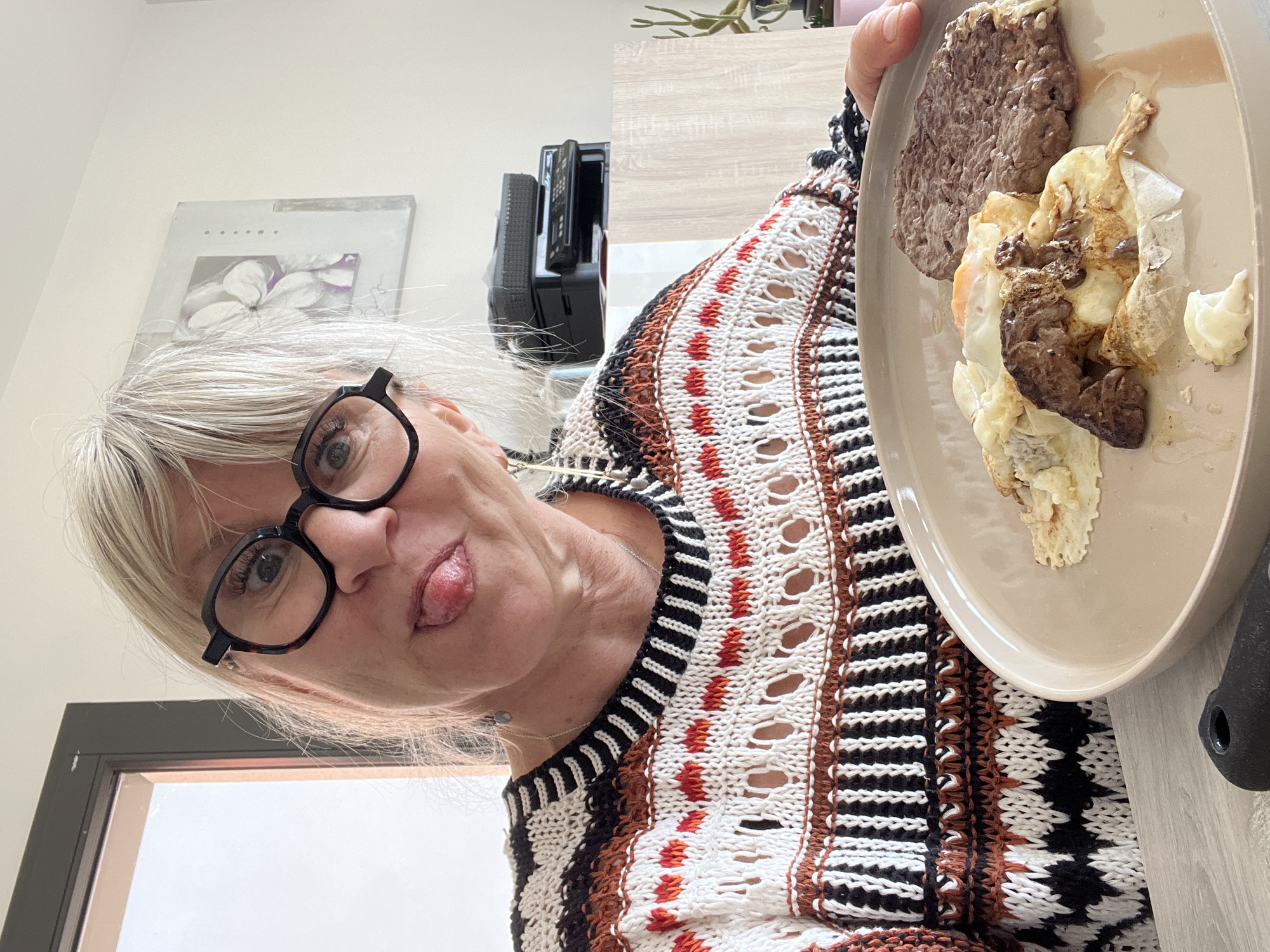 A woman with short blonde hair, wearing black-rimmed glasses and a white, brown, and black patterned sweater, holds a tan plate of food towards the camera. The plate contains a brown slab resembling meat, a fried egg, and a dollop of what appears to be mashed potatoes or cream. The woman is sticking her tongue out at the camera, suggesting a playful or humorous attitude. Behind her are indistinct items on a shelf and a painting with a flower. The scene is indoors, with a door visible in the background. There is no information to confirm the location.